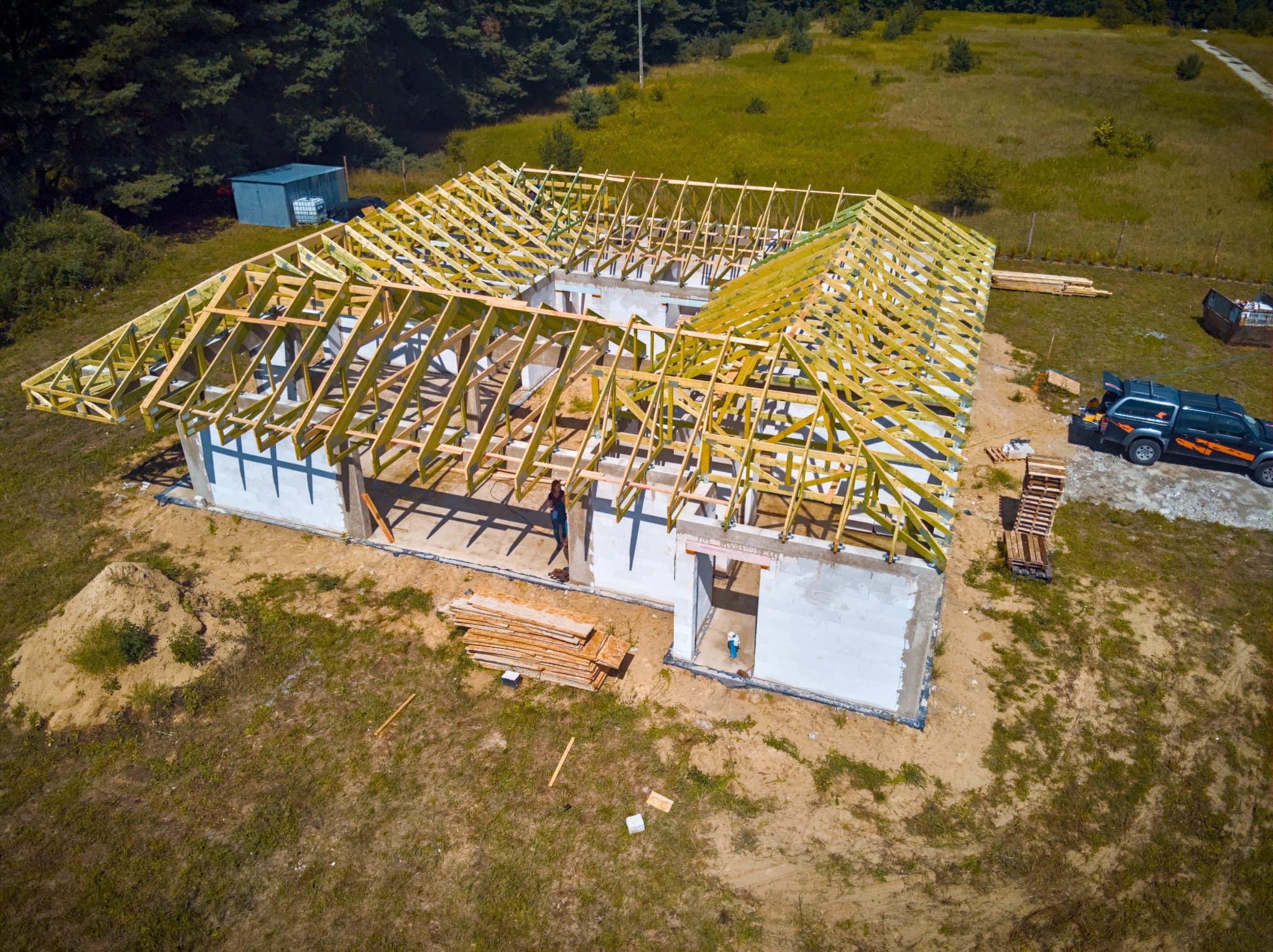 Widok z drona na budowę domu z częściowo wykonaną więźbą dachową, widoczne elementy konstrukcyjne z jasnego drewna, ściany z bloczków betonowych, teren budowy z piaskiem i trawą, w tle las...