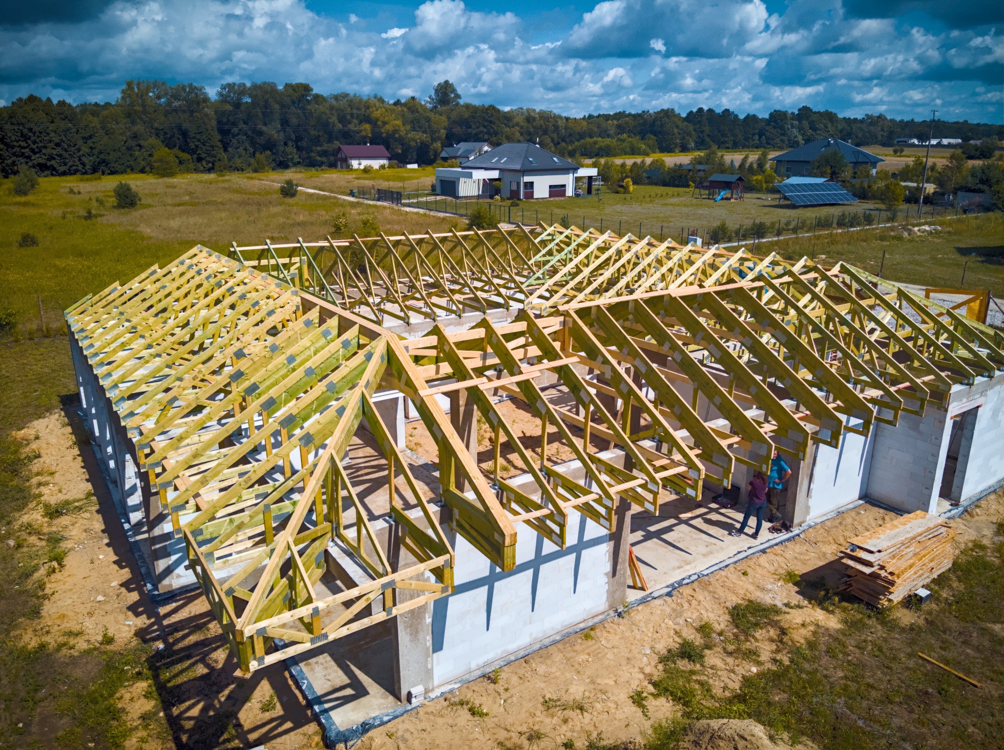 Wysoka perspektywa na konstrukcję dachu z drewnianych belek na budowie domu jednorodzinnego z białymi ścianami, w tle zielone pola i zabudowania.