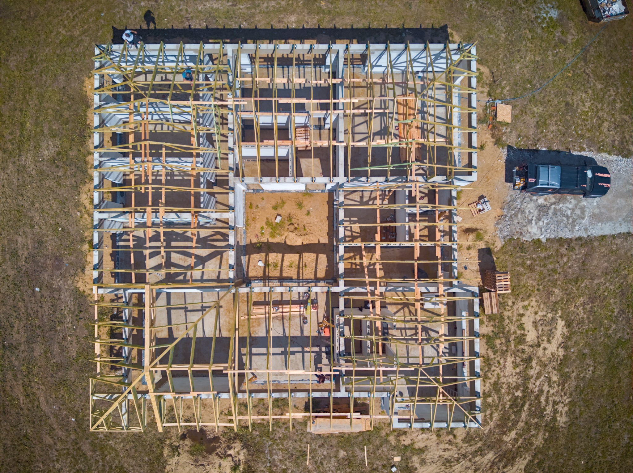 Widok z drona na fundamenty i drewnianą konstrukcję ścian powstającego domu, z zaparkowanym samochodem i widoczną osobą na placu budowy.