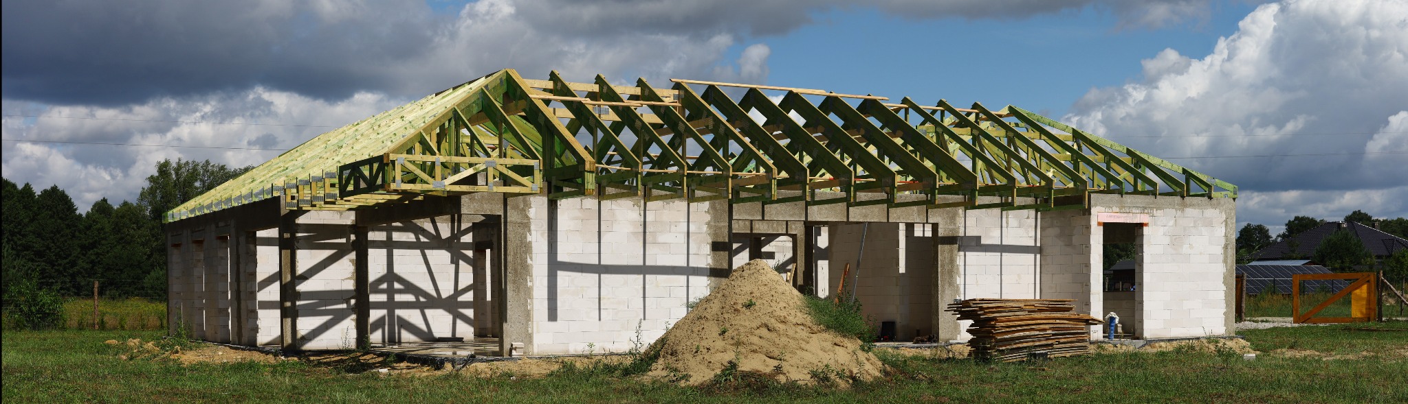 Budowa domu w stanie surowym otwartym z widoczną więźbą dachową, stertą piasku i deskami na trawie, w tle pochmurne niebo.