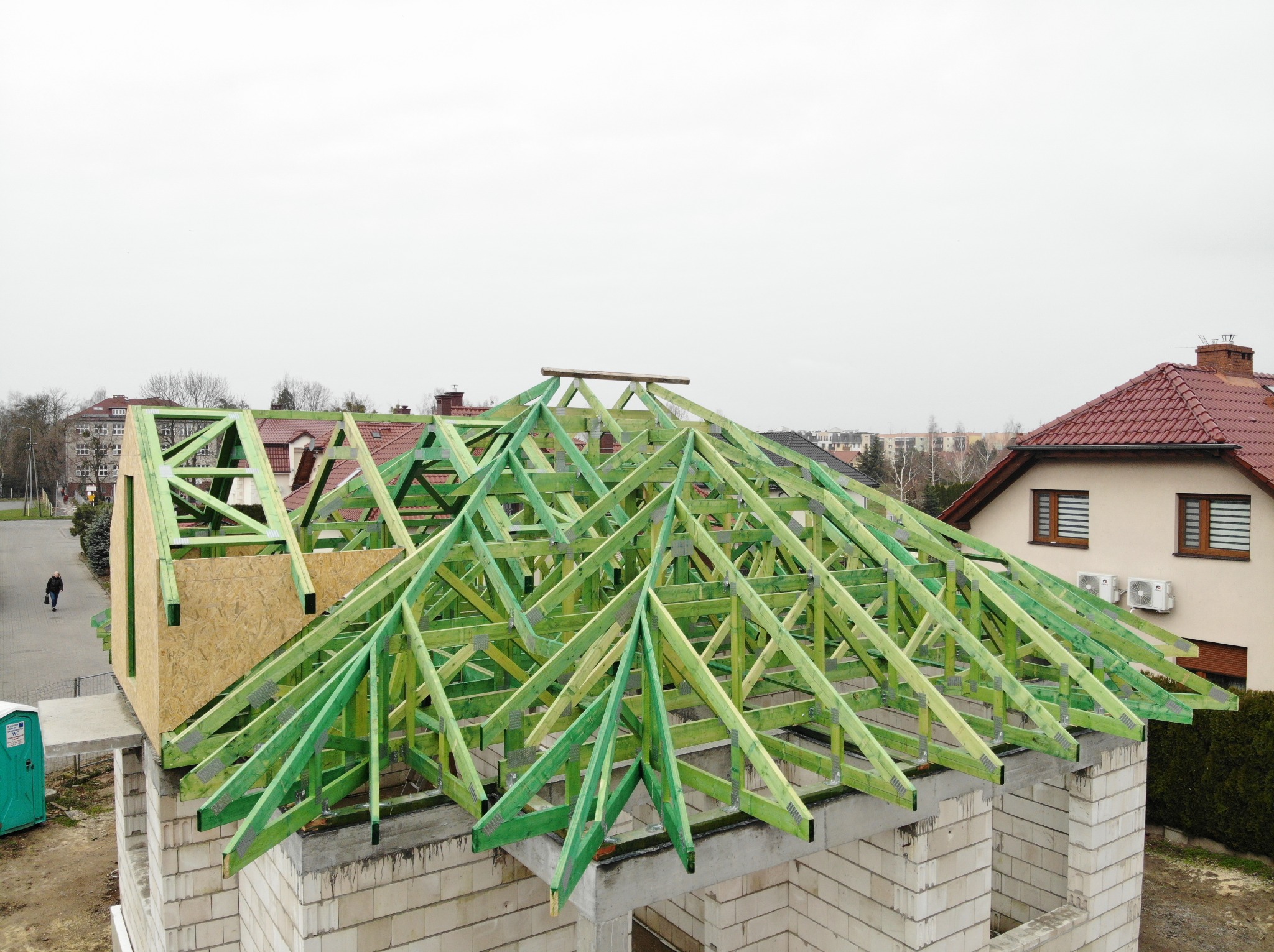 Wznoszenie konstrukcji dachu z zielonych belek na budynku z białych bloczków, widok z góry na osiedle domów jednorodzinnych w pochmurny dzień.
