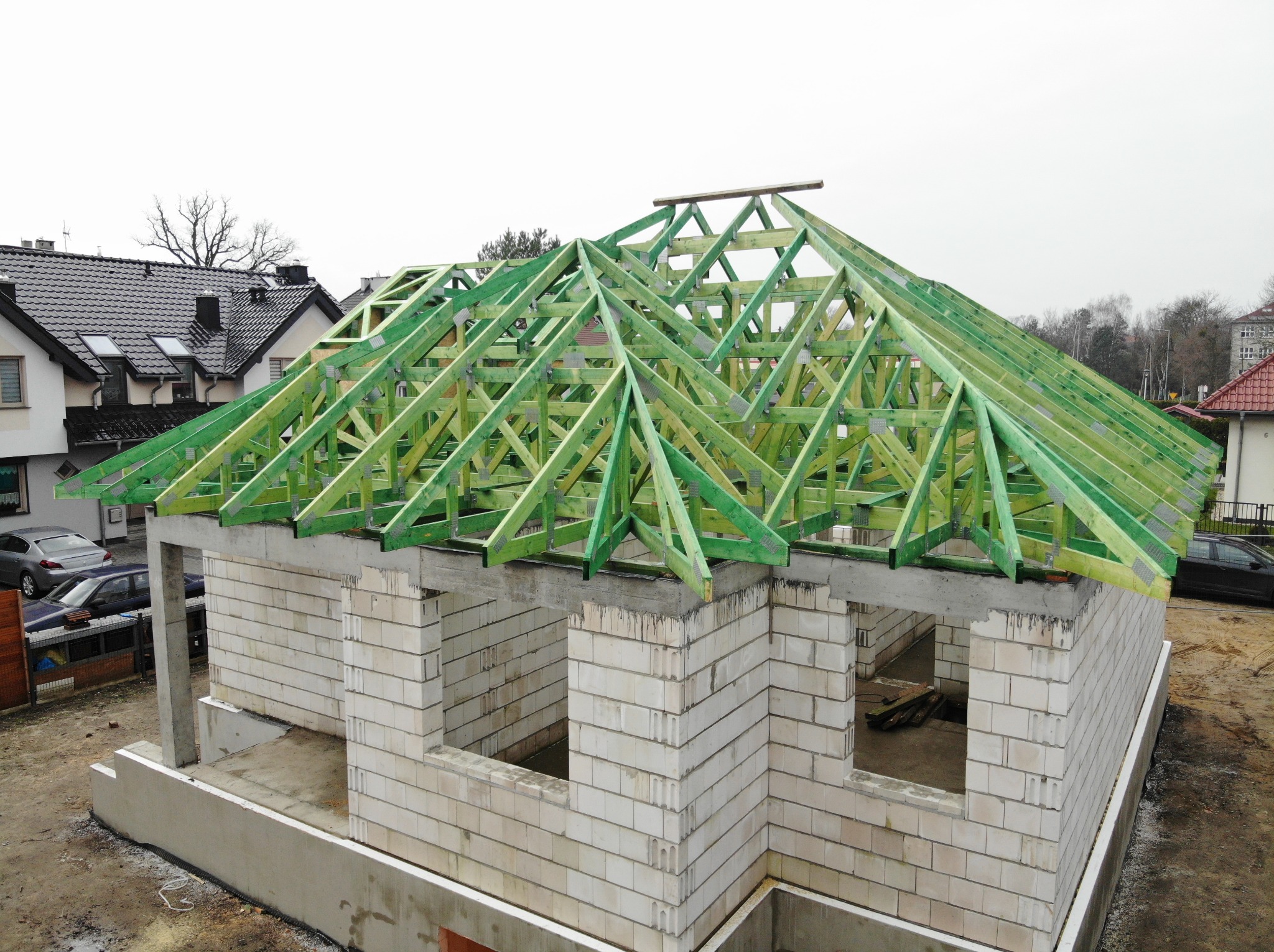 Surowy stan domu z białego bloczka, z zieloną drewnianą konstrukcją dachu w trakcie budowy, widok z góry na tle zabudowy jednorodzinnej i pochmurnego nieba.