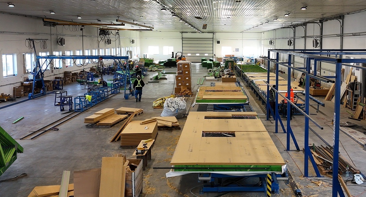 Wnętrze hali produkcyjnej z widocznymi elementami do budowy domów szkieletowych lub modułowych, w tym gotowe ściany z wycięciami na okna na linii produkcyjnej, pracownik w kasku i kamizelce...