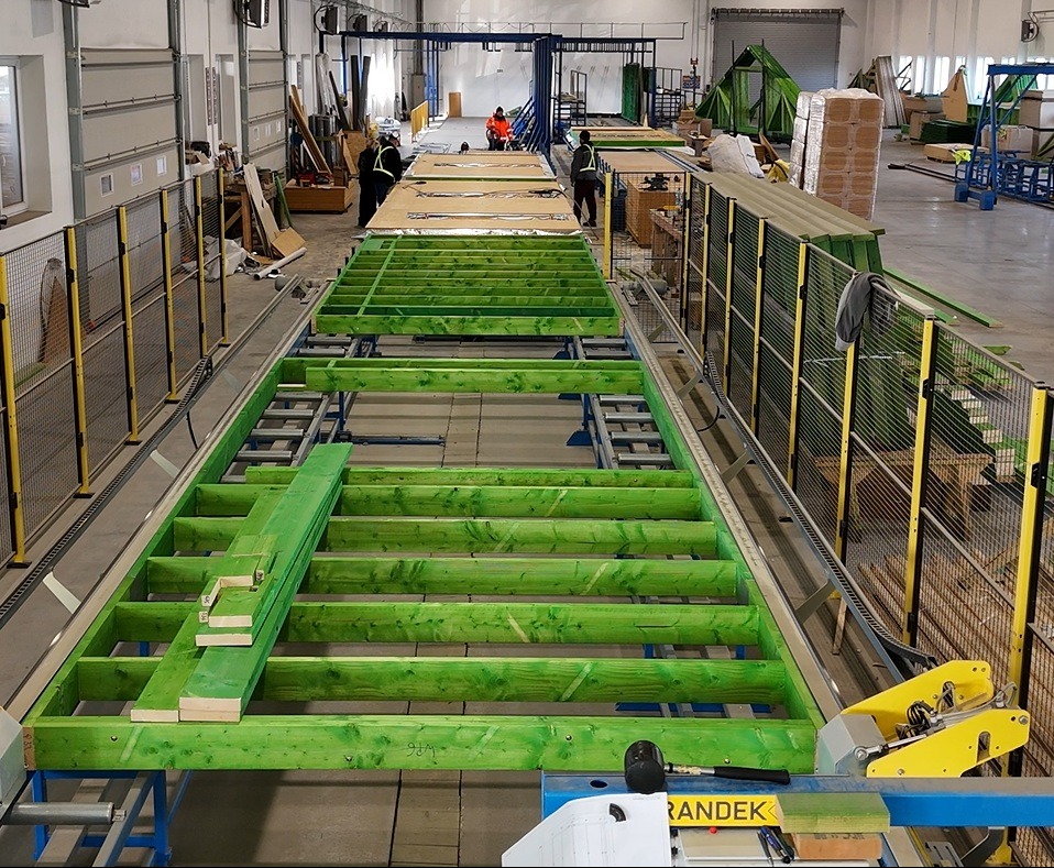 Produkcja zielonej ramy podłogowej w hali w Wiechlicach, widoczni pracownicy w oddali i maszyny do obróbki drewna.