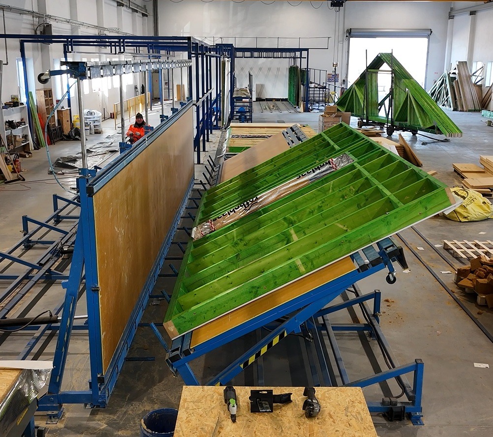 Produkcja elementów konstrukcyjnych: ściana z płyty OSB w pionowej ramie i zielona rama dachu na ruchomej platformie, w tle pracownik w pomarańczowej kurtce i gotowa konstrukcja dachu na wózku