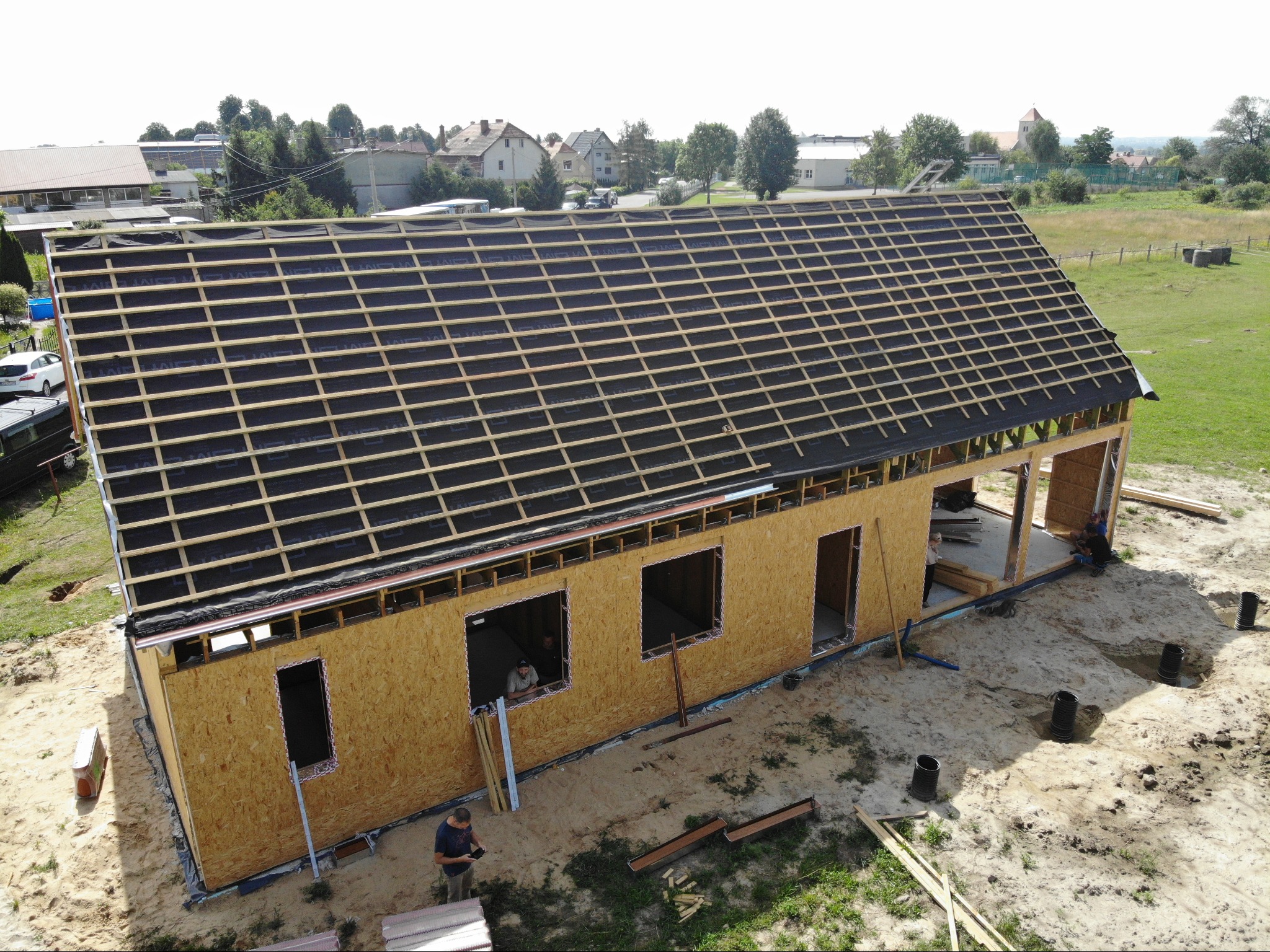 Budowa domu w stanie surowym otwartym: ściany z płyt OSB, widoczna więźba dachowa pokryta membraną, robotnicy na placu budowy, widok z góry.