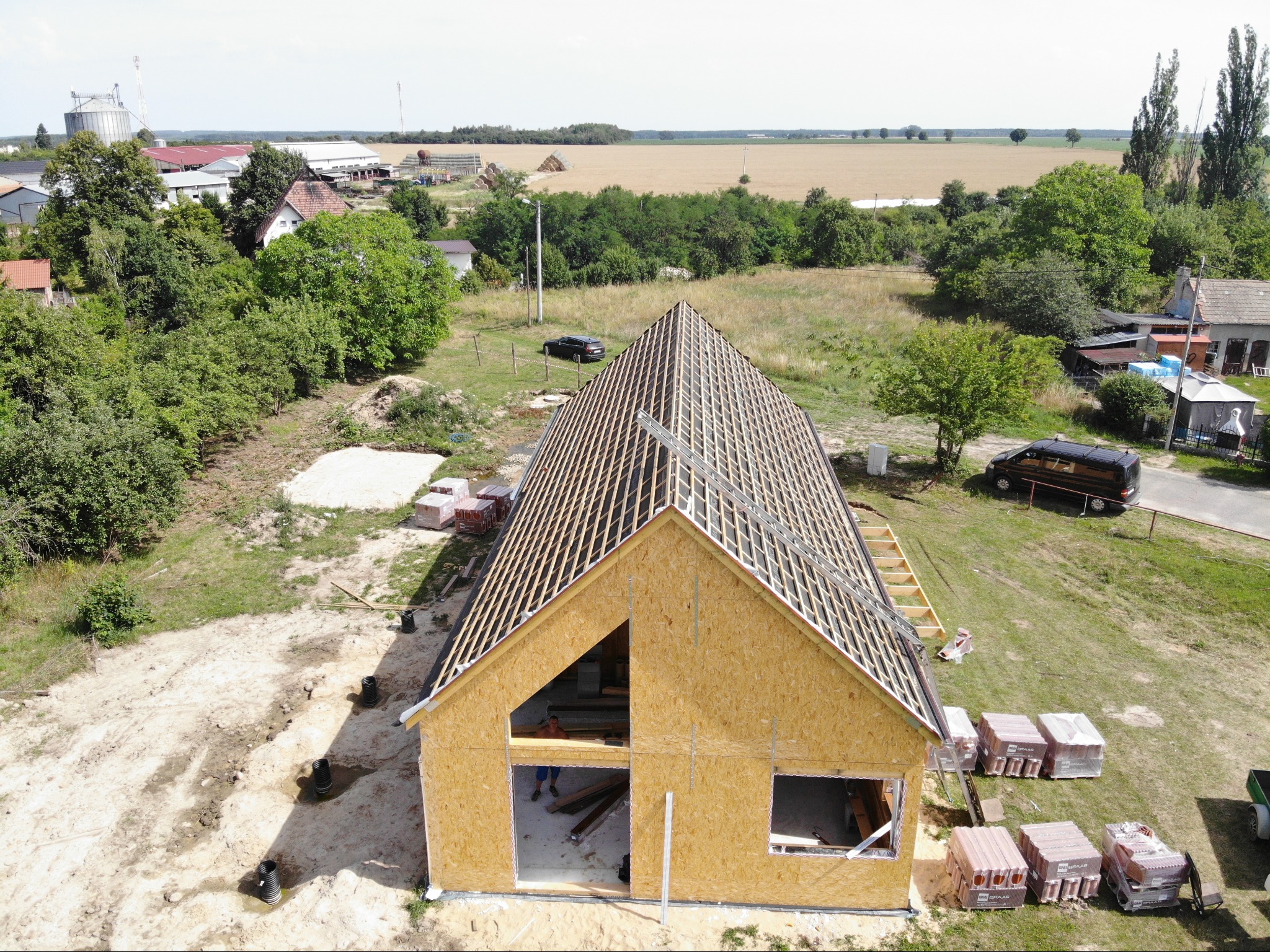 Wznoszenie konstrukcji budynku z drewnianym szkieletem i widoczną więźbą dachową, ujęcie z góry na tle wiejskiego krajobrazu z polami i zabudowaniami.