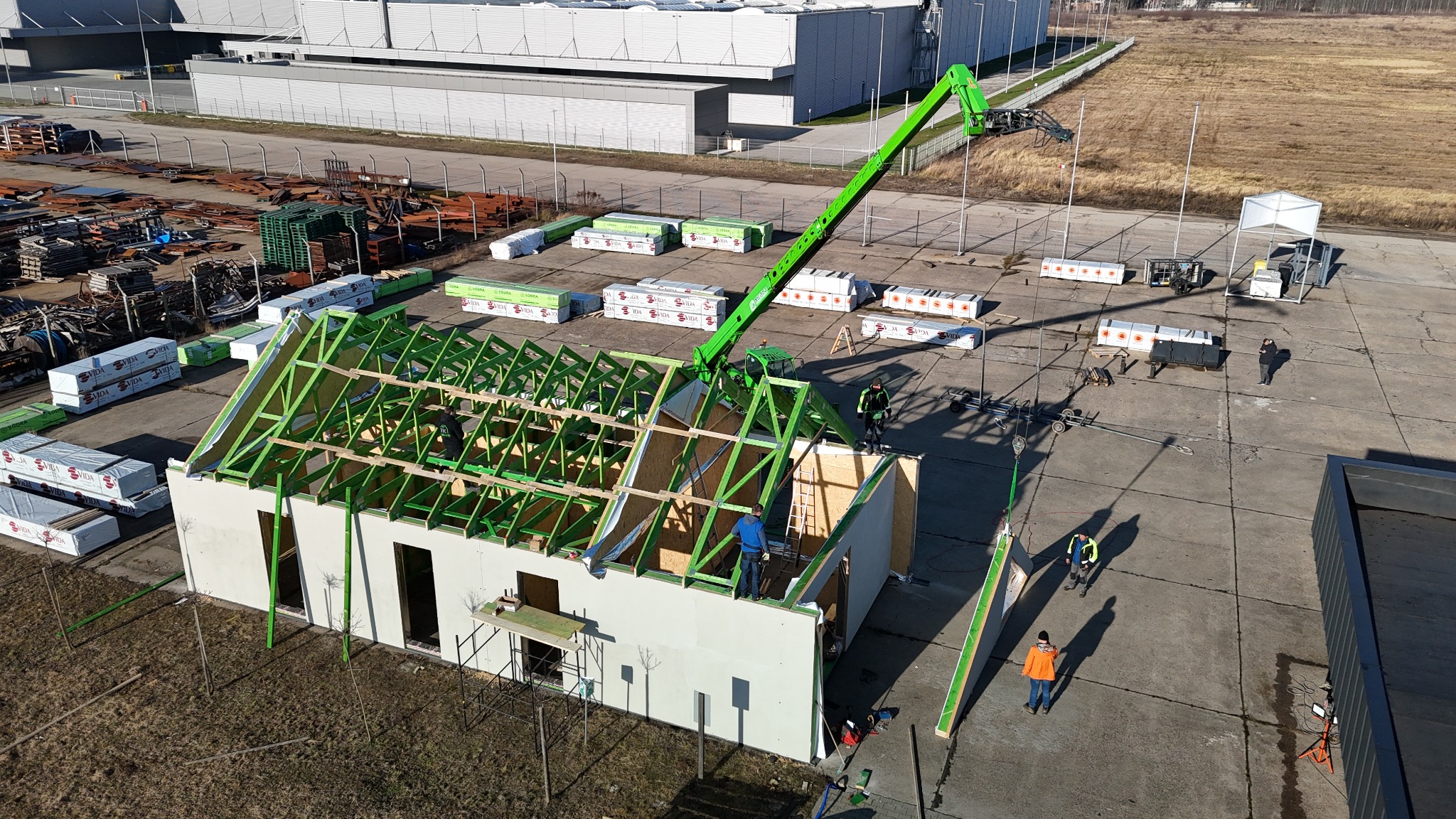 Montaż konstrukcji domu z prefabrykatów na betonowej płycie, widoczny dźwig i pracownicy podczas pracy na wysokościach.