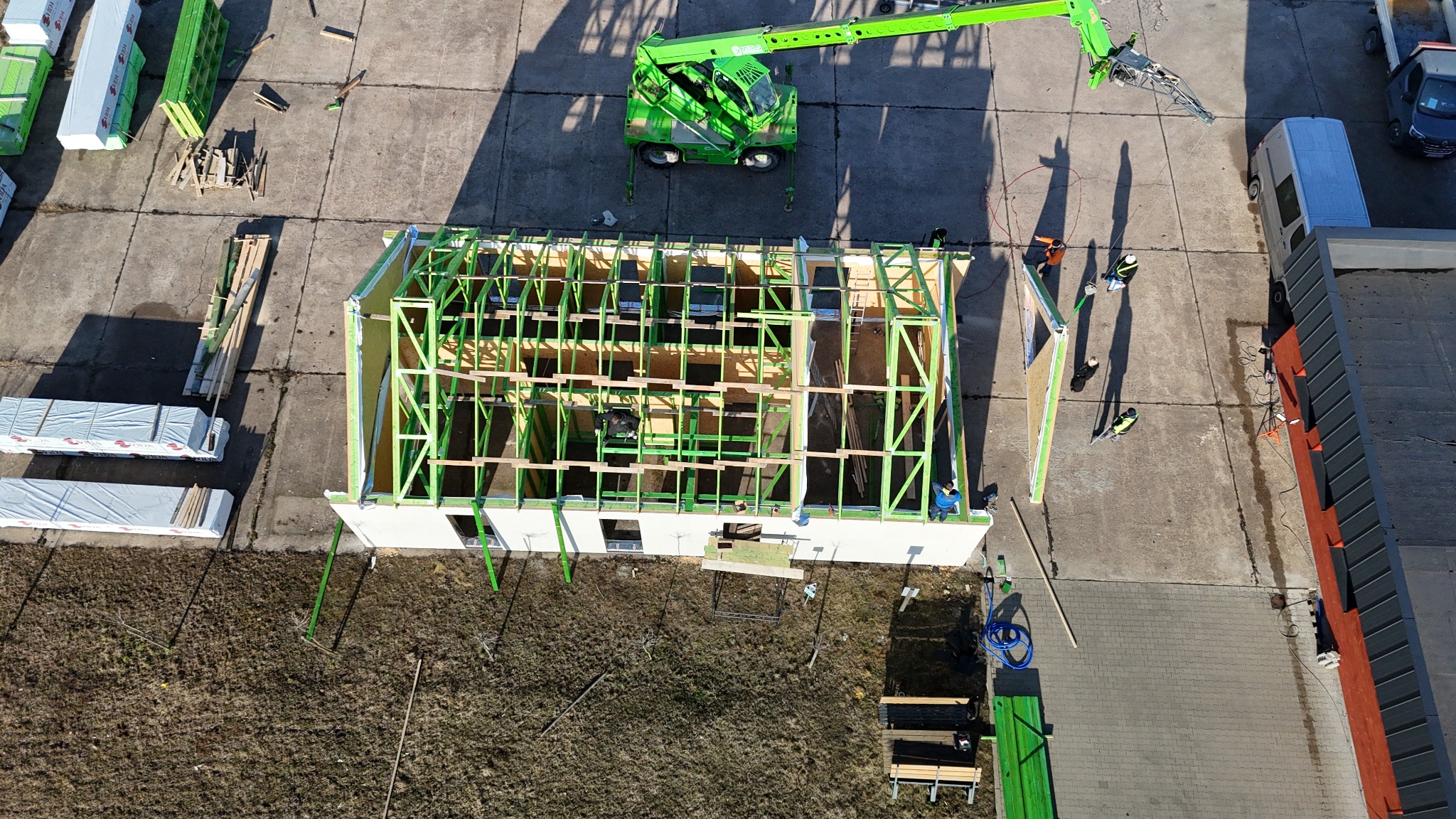 Budowa domu szkieletowego z zieloną konstrukcją, widok z góry z dźwigiem i pracownikami w kaskach, materiały budowlane obok