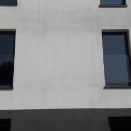 PLANETA SZKŁA SPÓŁKA Z OGRANICZONĄ ODPOWIEDZIALNOŚCIĄ - Elewacja budynku z dwoma prostokątnymi oknami z szarymi ramami i szkłem refleksyjnym, widoczne odbicie drzewa w jednym z okien.