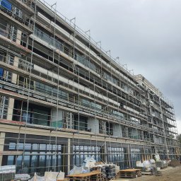 PLANETA SZKŁA SPÓŁKA Z OGRANICZONĄ ODPOWIEDZIALNOŚCIĄ - Wielopiętrowy budynek w trakcie montażu dużych okien, otoczony rusztowaniami, z widocznymi paletami i materiałami budowlanymi na placu budowy.