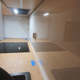 PLANETA SZKŁA SPÓŁKA Z OGRANICZONĄ ODPOWIEDZIALNOŚCIĄ - Szklana ściana nad blatem kuchennym z zamontowanymi płytami indukcyjnymi, zabezpieczona niebieską taśmą malarską, widok na wnętrze kuchni z drewnianymi szafkami.