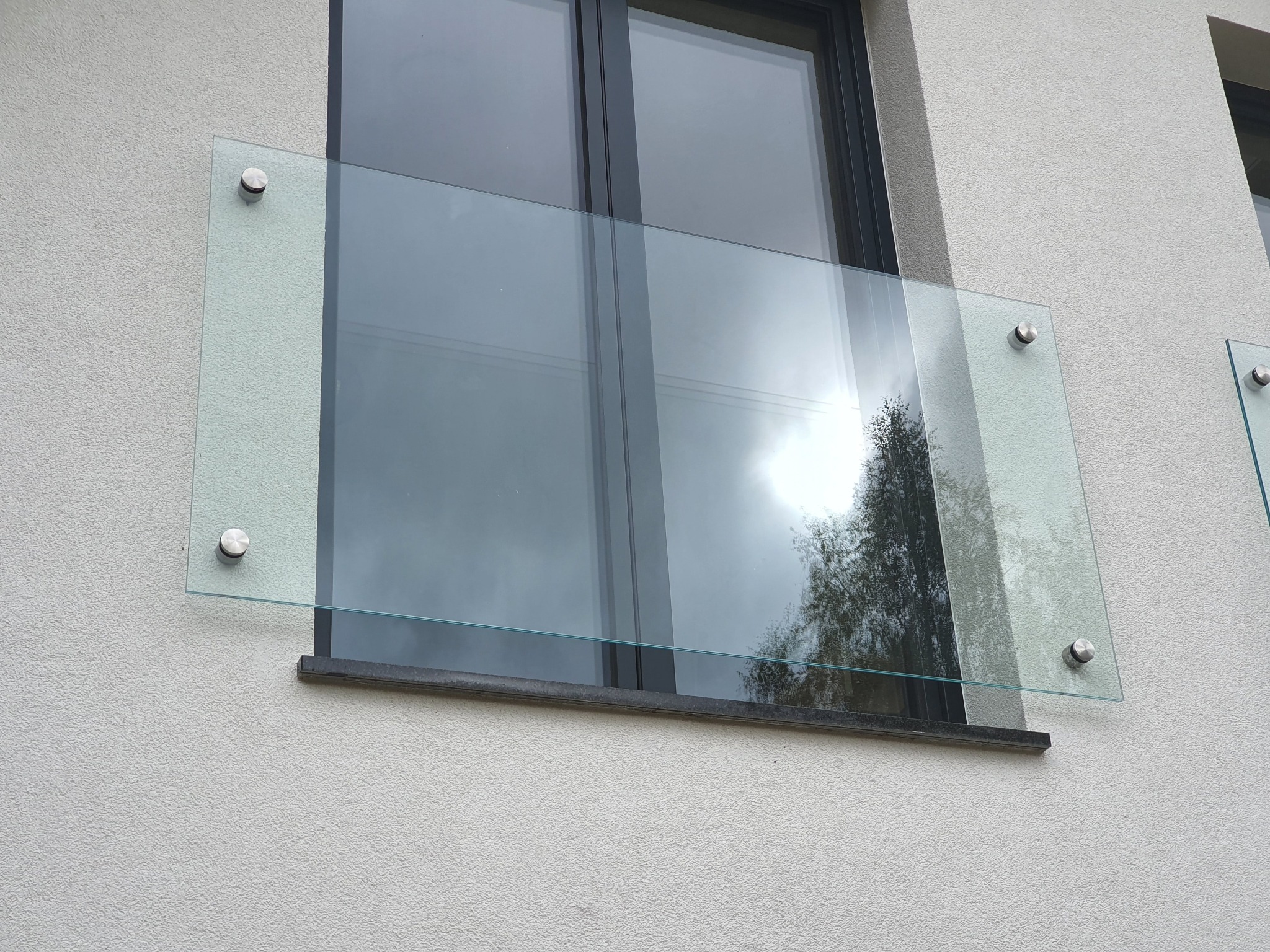 Szklana osłona okienna zamontowana na elewacji budynku za pomocą metalowych uchwytów punktowych, odbijająca w szybie drzewo i niebo.