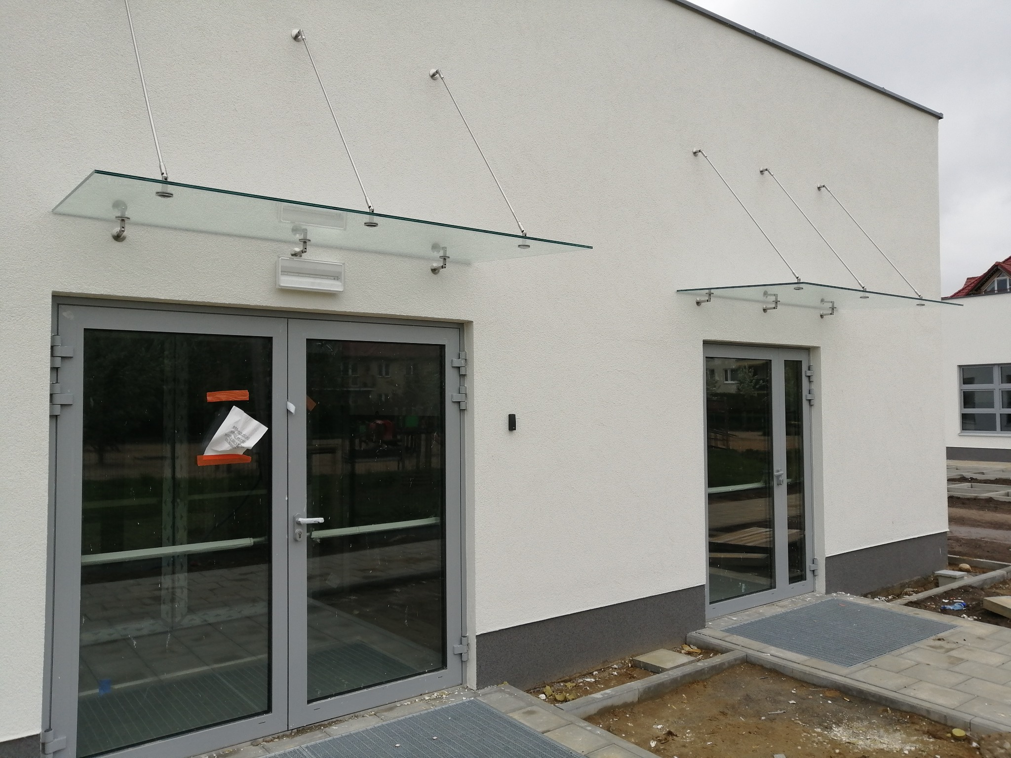 Dwa wejścia do budynku z szarymi, przeszklonymi drzwiami i szklanymi daszkami wspartymi na metalowych prętach, widoczny fragment placu budowy.