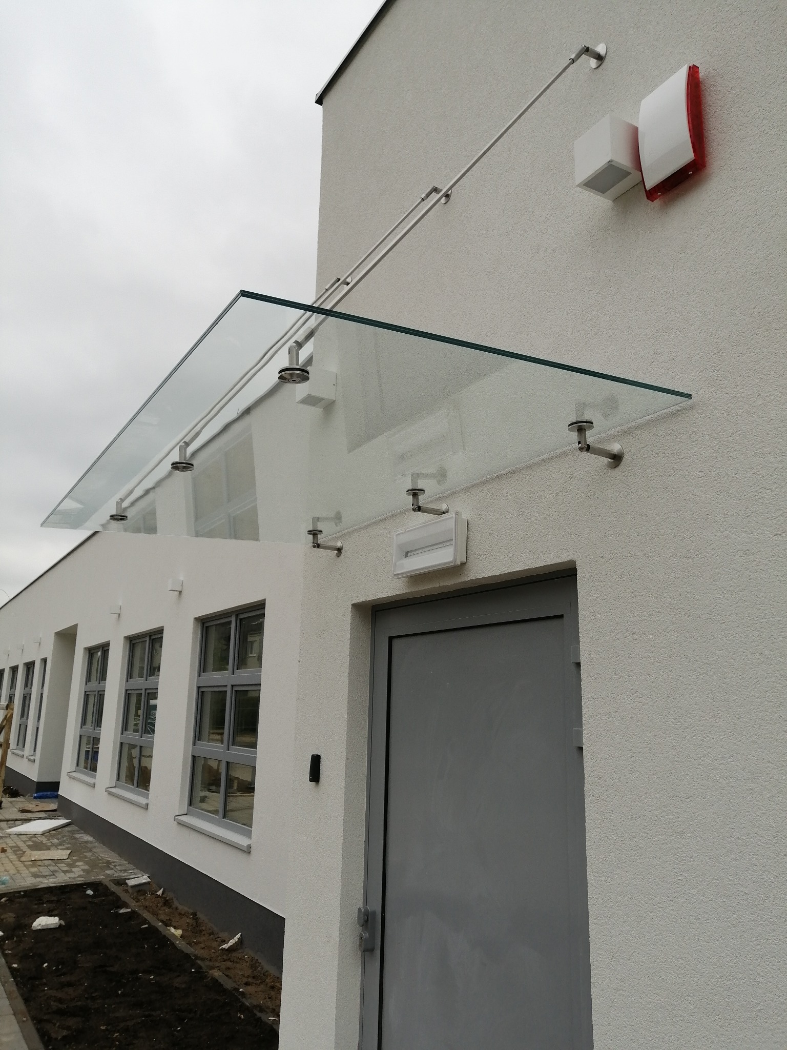 Szklany daszek nad wejściem do budynku z szarą elewacją, zamontowany na metalowych wspornikach, widoczny system alarmowy.