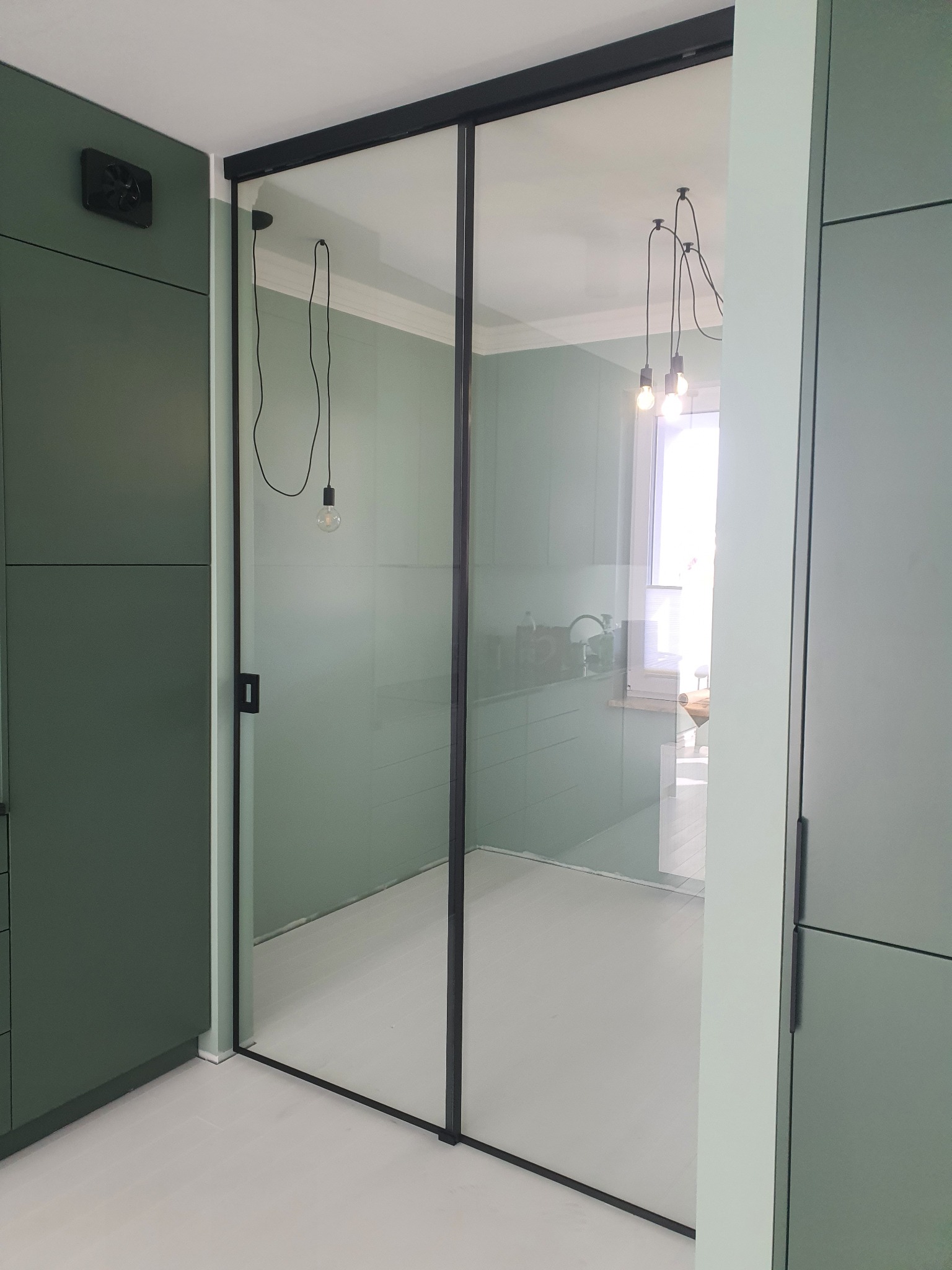 Przesuwne drzwi szklane z czarną ramą, widok na wnętrze łazienki z białymi podłogami i wiszącymi lampami z żarówkami Edisona.