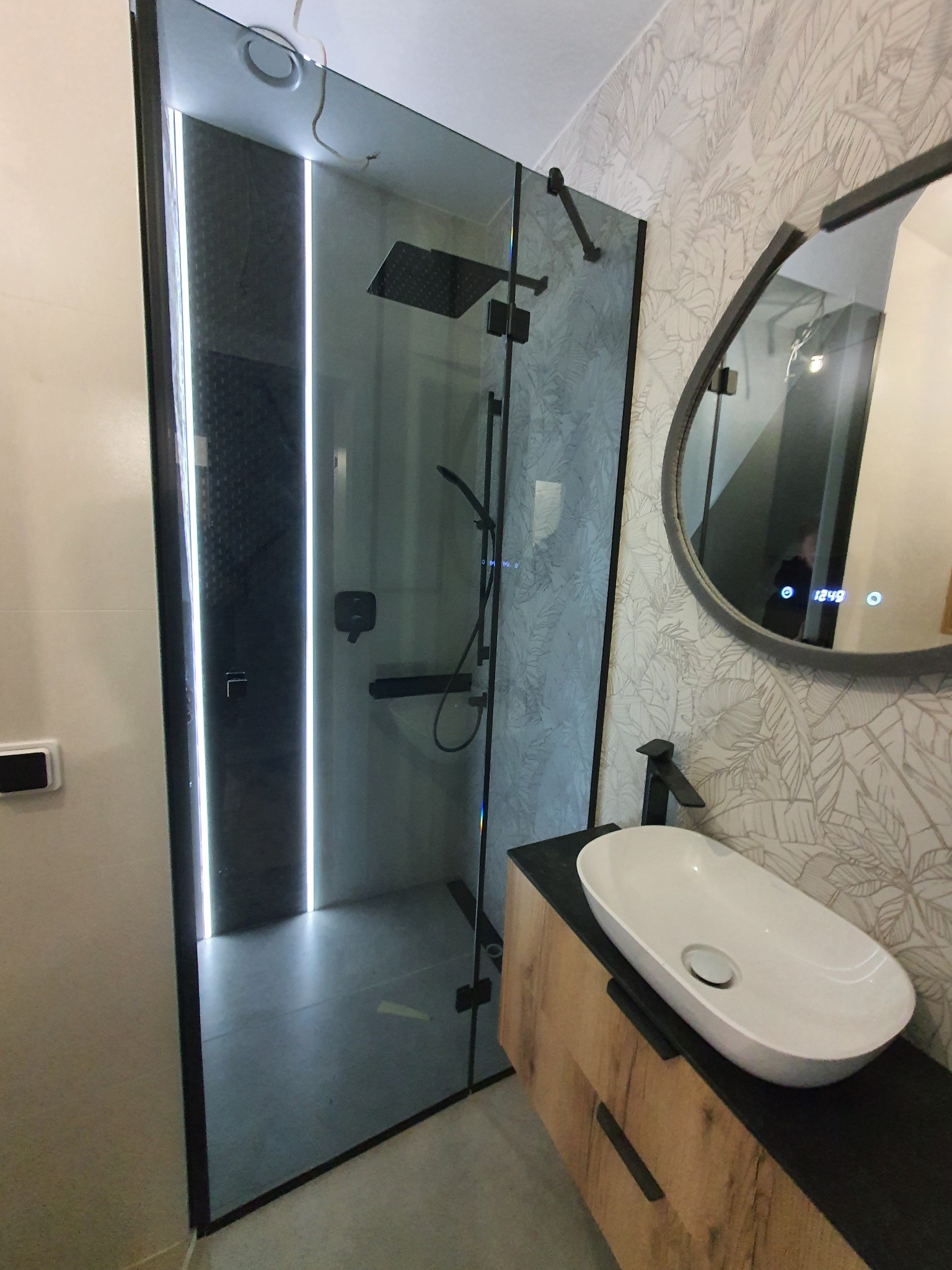 Nowoczesna kabina prysznicowa ze szklaną ścianą z podświetleniem LED, widok na umywalkę nablatową i lustro z wyświetlaczem cyfrowym w łazience z tapetą w liście.