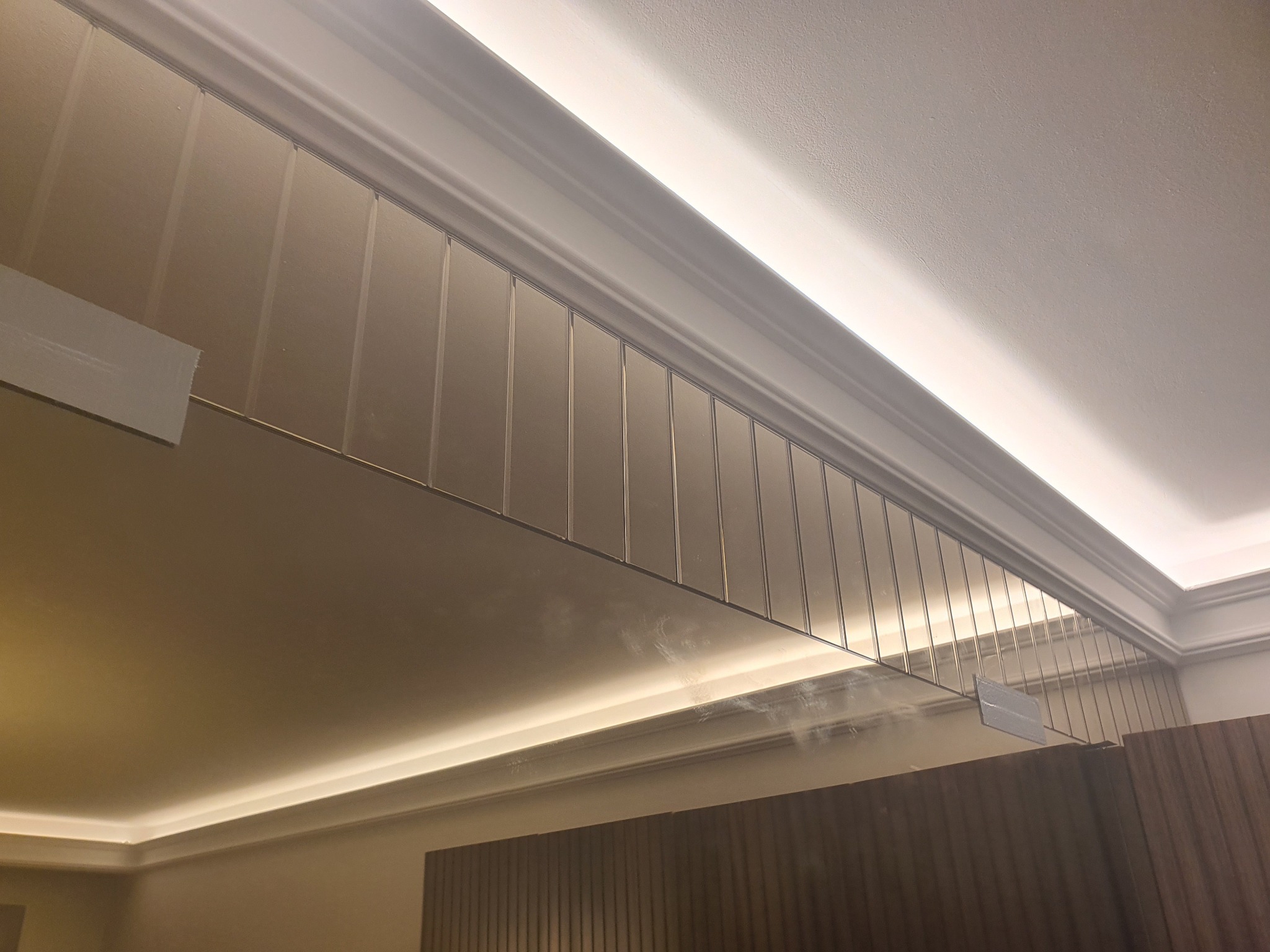 Szklana tafla zamontowana pod sufitem z oświetleniem LED, widoczne elementy mocujące i odbicie drewnianej ściany.