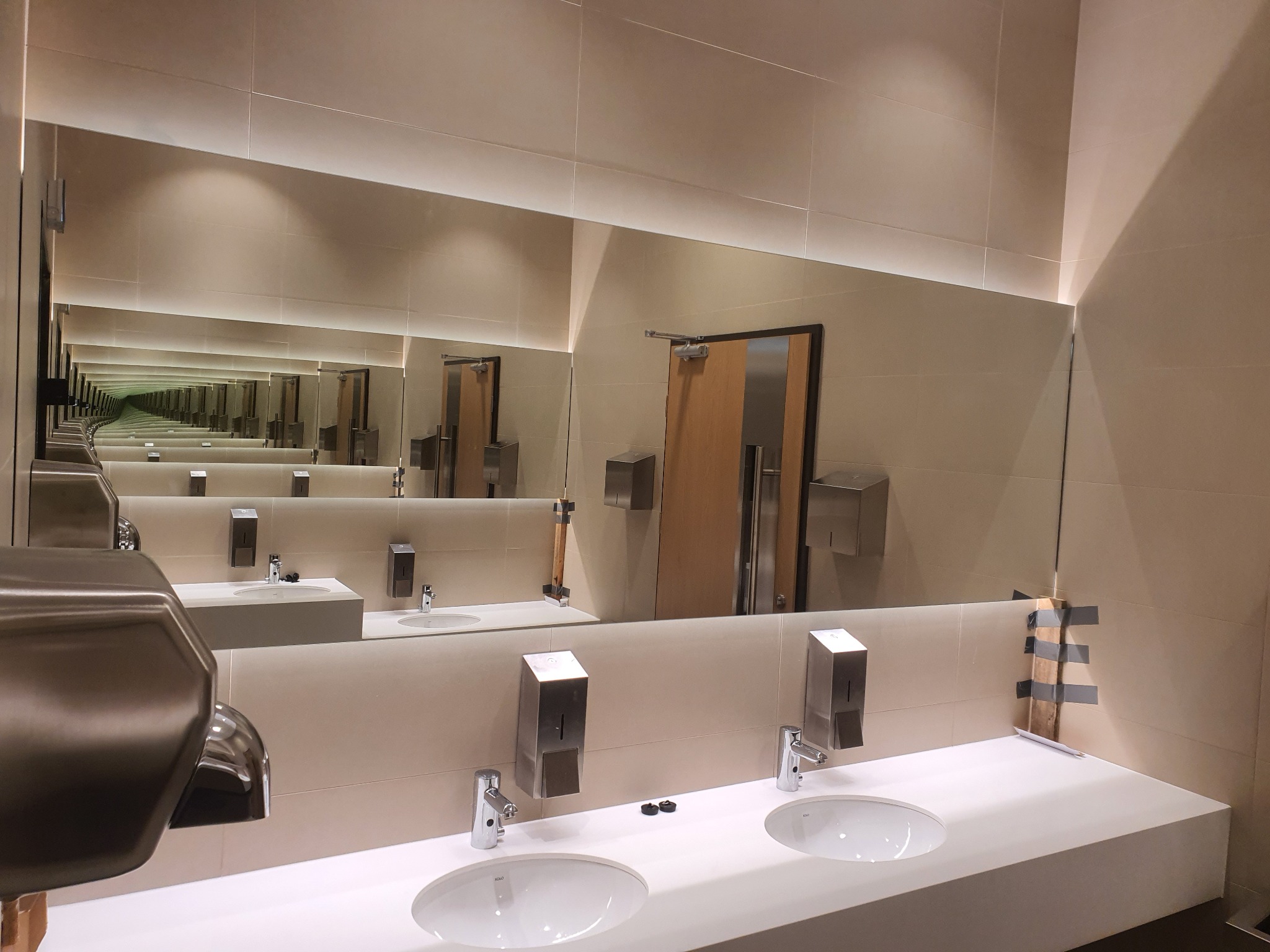 Długa, pozioma tafla lustra w publicznej toalecie, tworząca efekt nieskończoności odbić umywalek i dozowników mydła. Widoczne jasne płytki ceramiczne i oświetlenie LED.