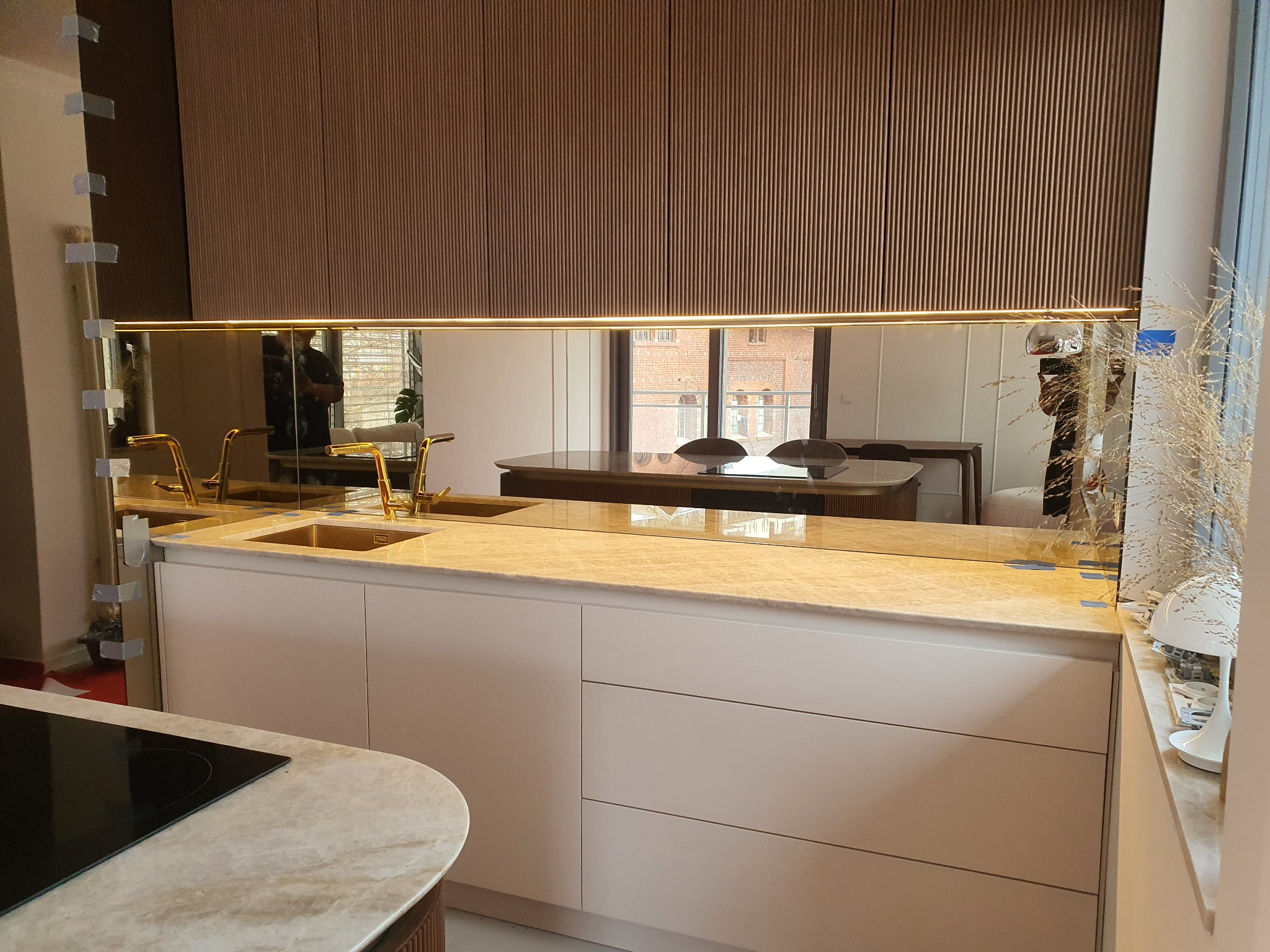 Kuchnia z beżowymi frontami szafek, marmurowym blatem i lustrzanym panelem na ścianie, odbijającym jadalnię. Na lustrze widoczne taśmy montażowe.