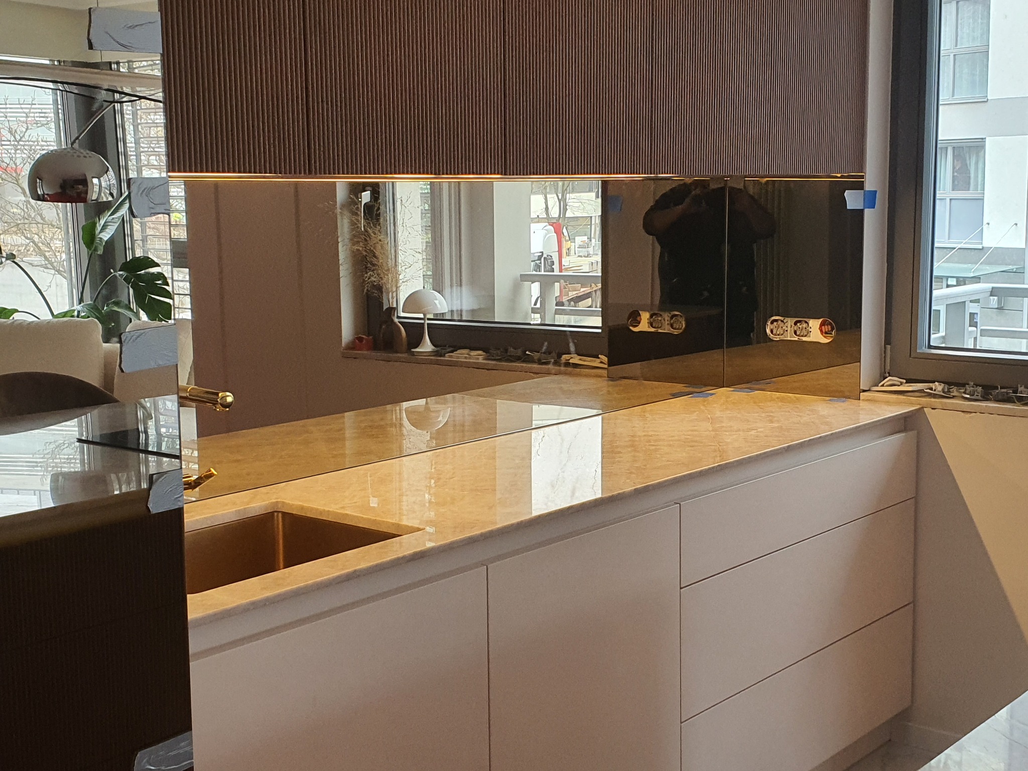 Szklany panel ścienny w kuchni, odbijający fragment okna i lampy, z widocznymi gniazdkami elektrycznymi i marmurowym blatem.