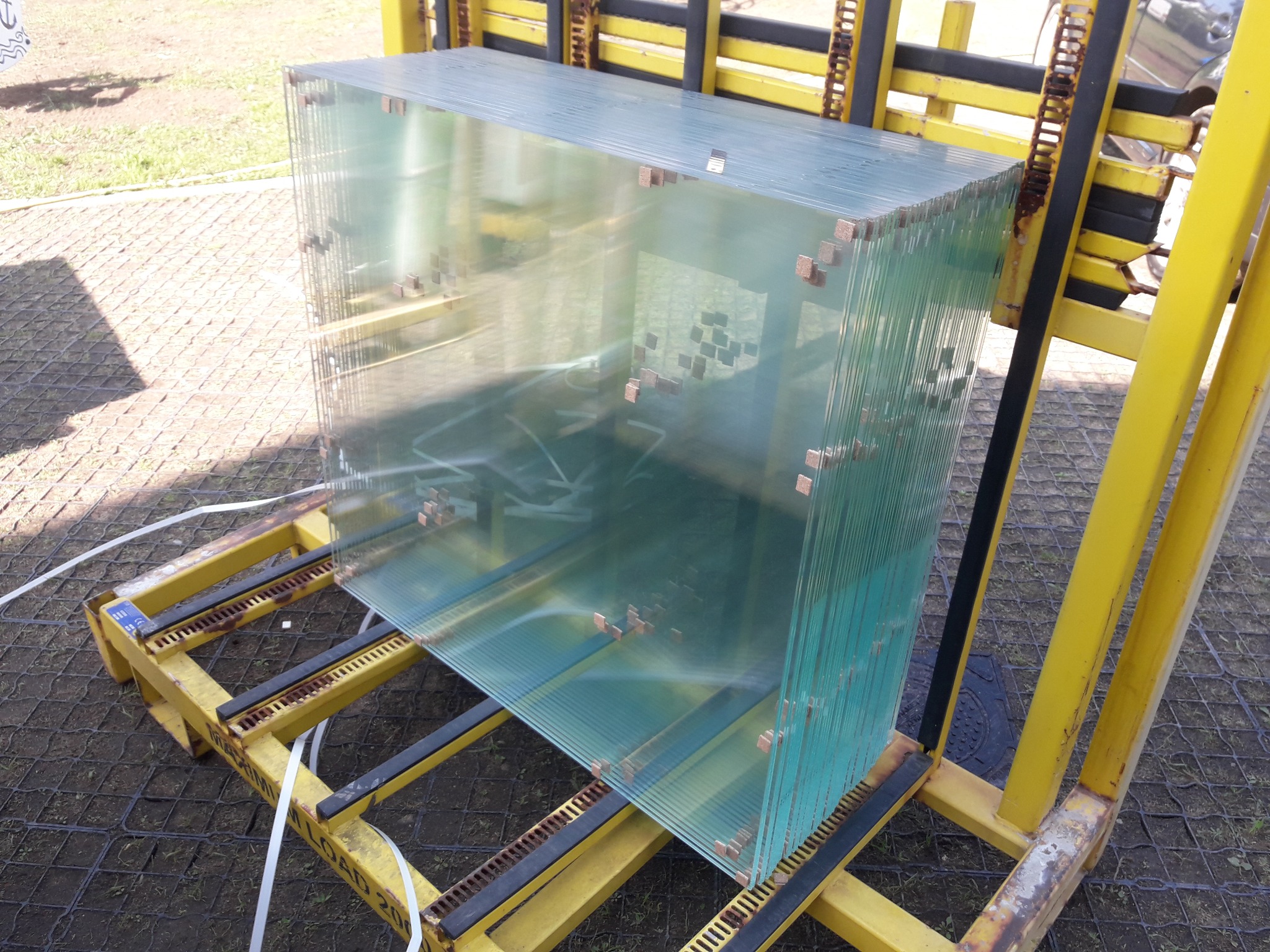 Stos tafli szkła zabezpieczonych przekładkami, umieszczonych na żółtym metalowym stojaku transportowym na zewnątrz.