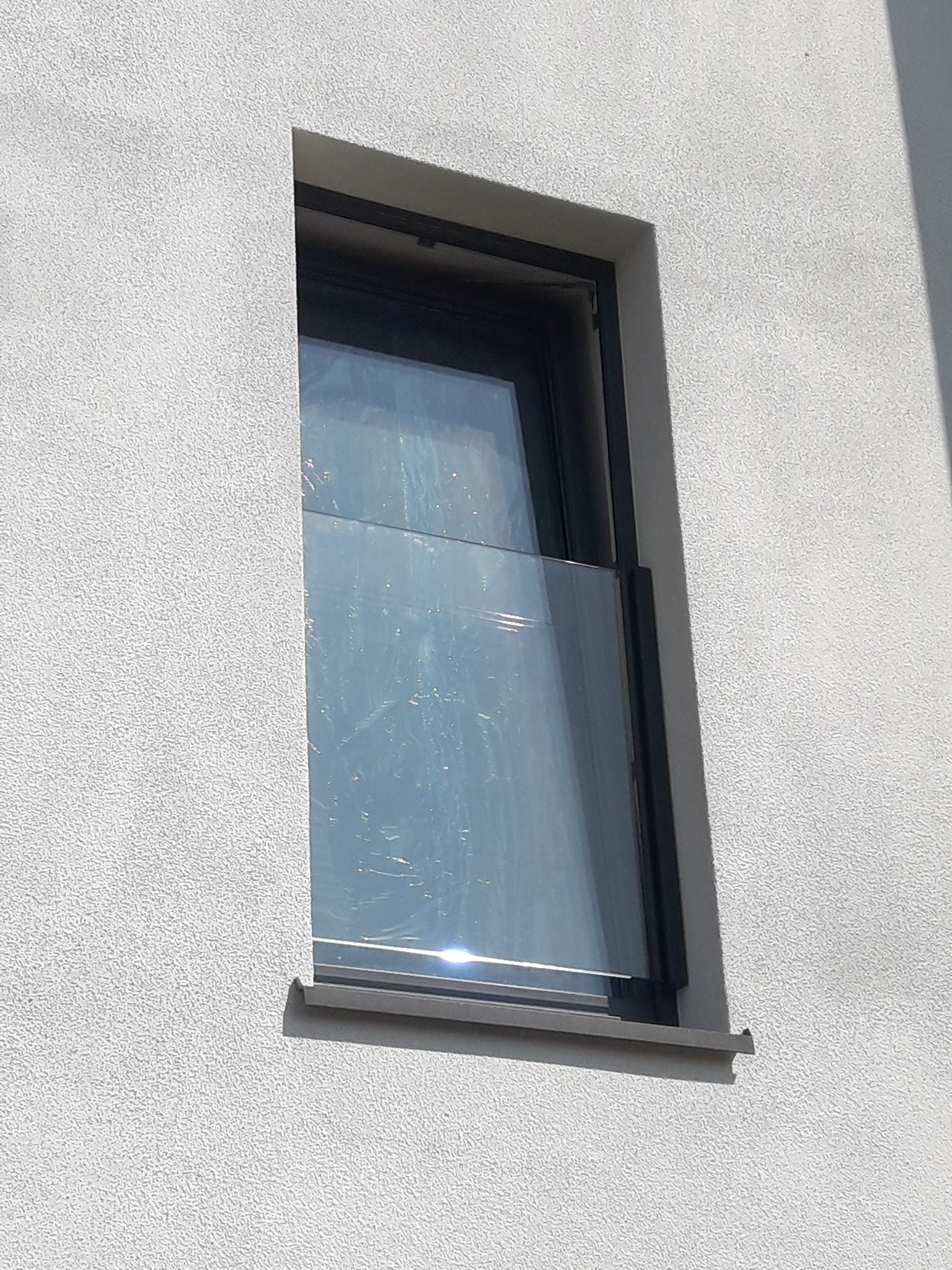 Nowo zamontowane okno z czarną ramą, zabezpieczone folią ochronną, w elewacji budynku o jasnej, chropowatej fakturze.