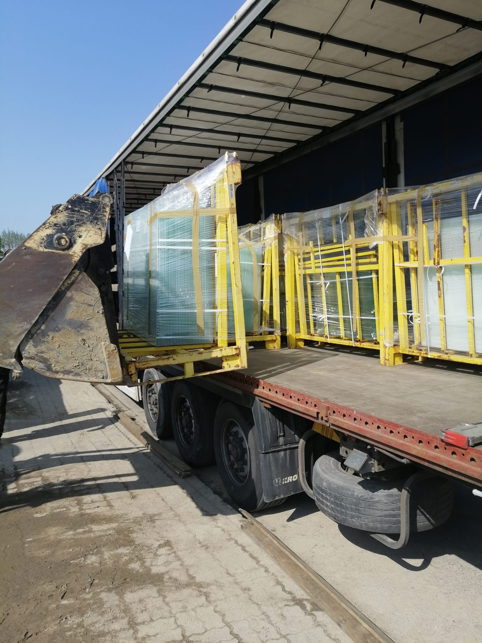 Załadunek pakietów szkła w żółtych ramach na naczepę ciężarówki przy użyciu wózka widłowego.