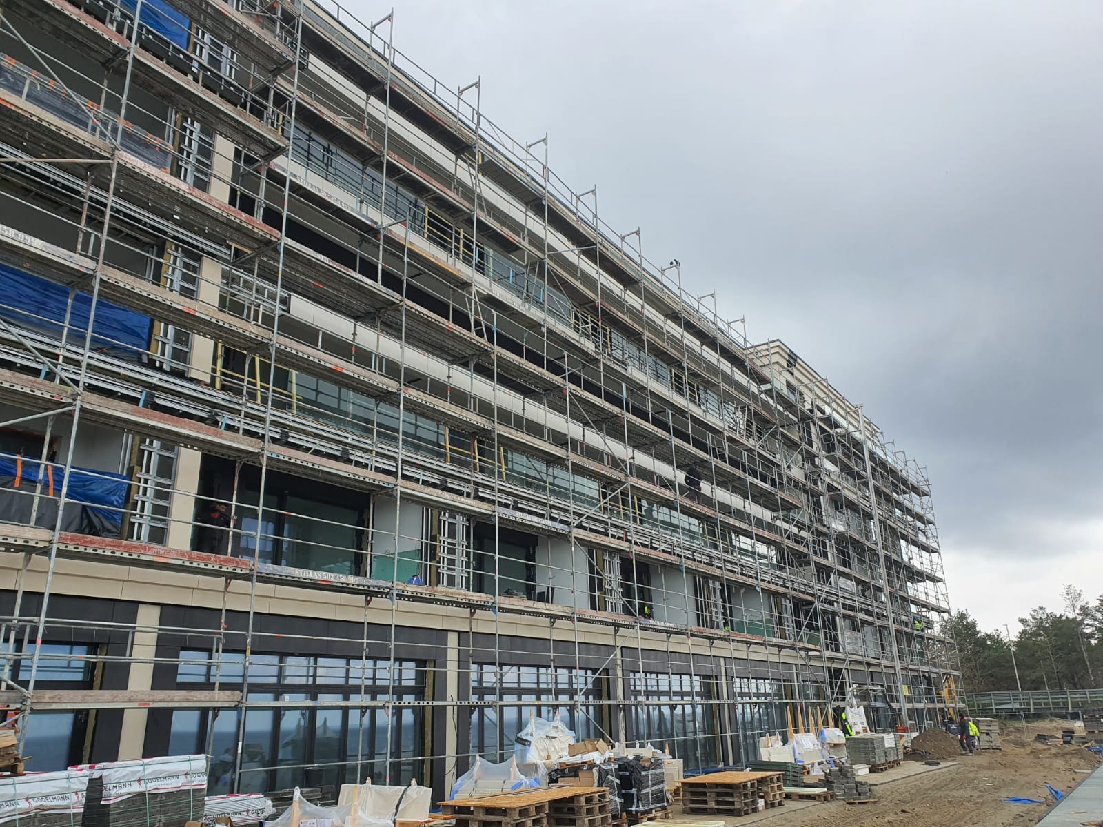 Wielopiętrowy budynek w trakcie montażu dużych okien, otoczony rusztowaniami, z widocznymi paletami i materiałami budowlanymi na placu budowy.