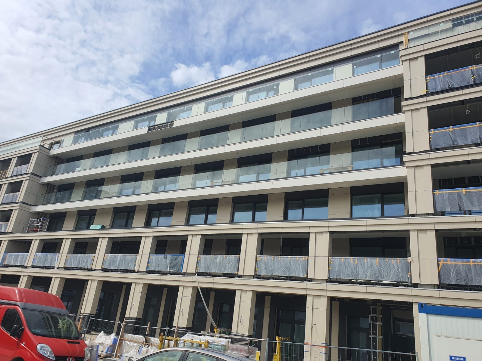 Wielopiętrowy budynek z przeszklonymi balkonami, niektóre z folią ochronną, widoczne samochody na pierwszym planie