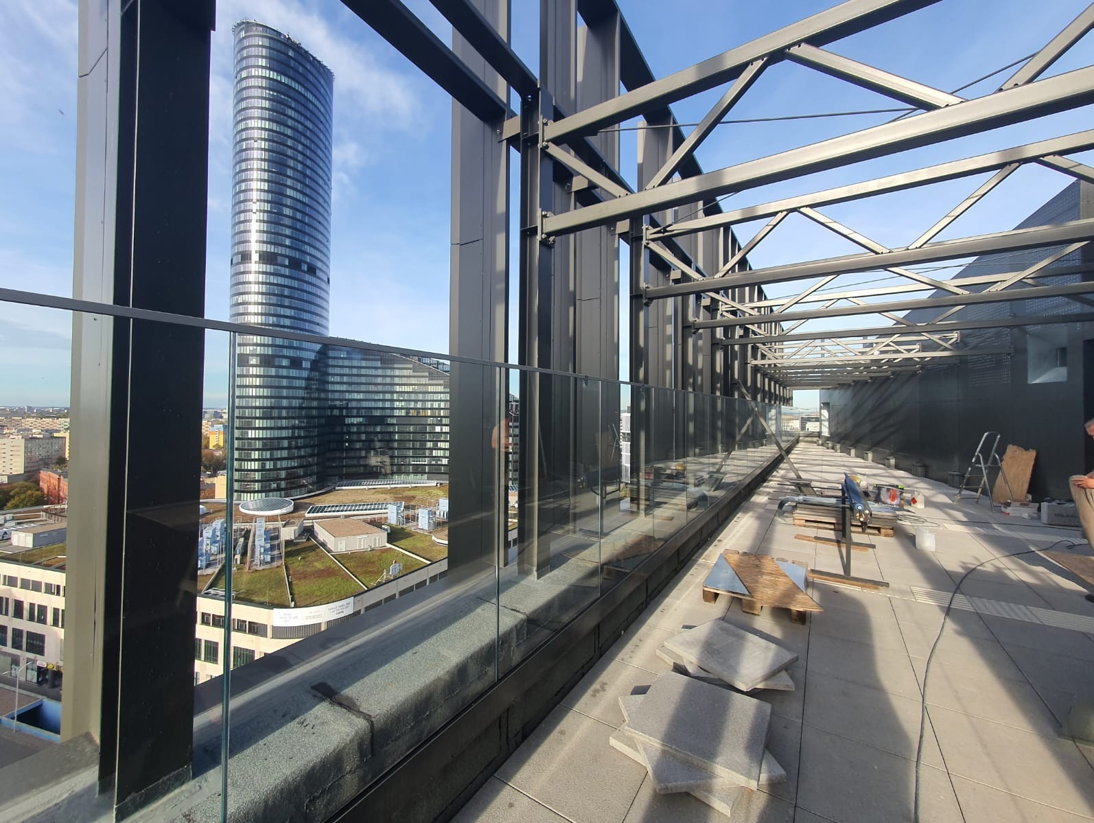 Szklana balustrada na tarasie budynku w trakcie montażu, widok na panoramę miasta z wysokim wieżowcem w tle.