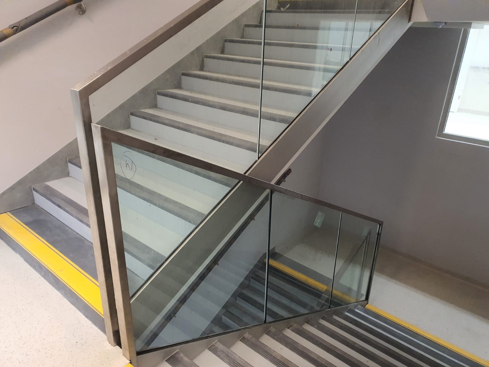 Szklana balustrada zabezpiecza schody w budynku w Warszawie, widoczne żółte pasy antypoślizgowe na stopniach, ujęcie z góry.