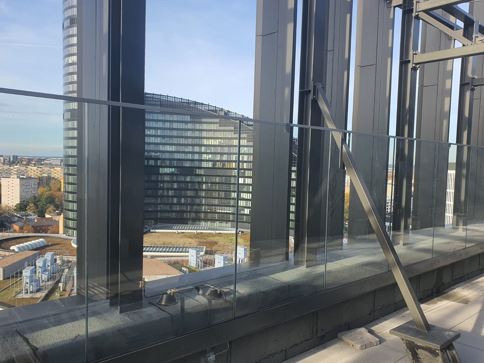 Szklana balustrada na tarasie wysokiego budynku z widokiem na panoramę miasta i inne wieżowce w słoneczny dzień.