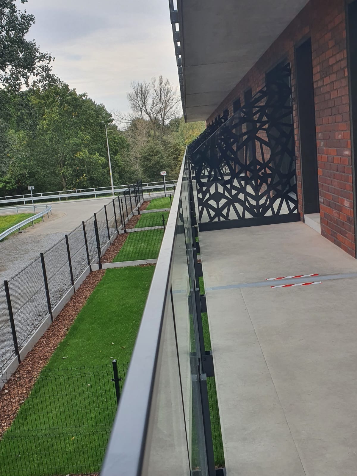 Balkon z balustradą ze szkła i dekoracyjnymi panelami ażurowymi, widok z boku, na tle zieleni i drogi.