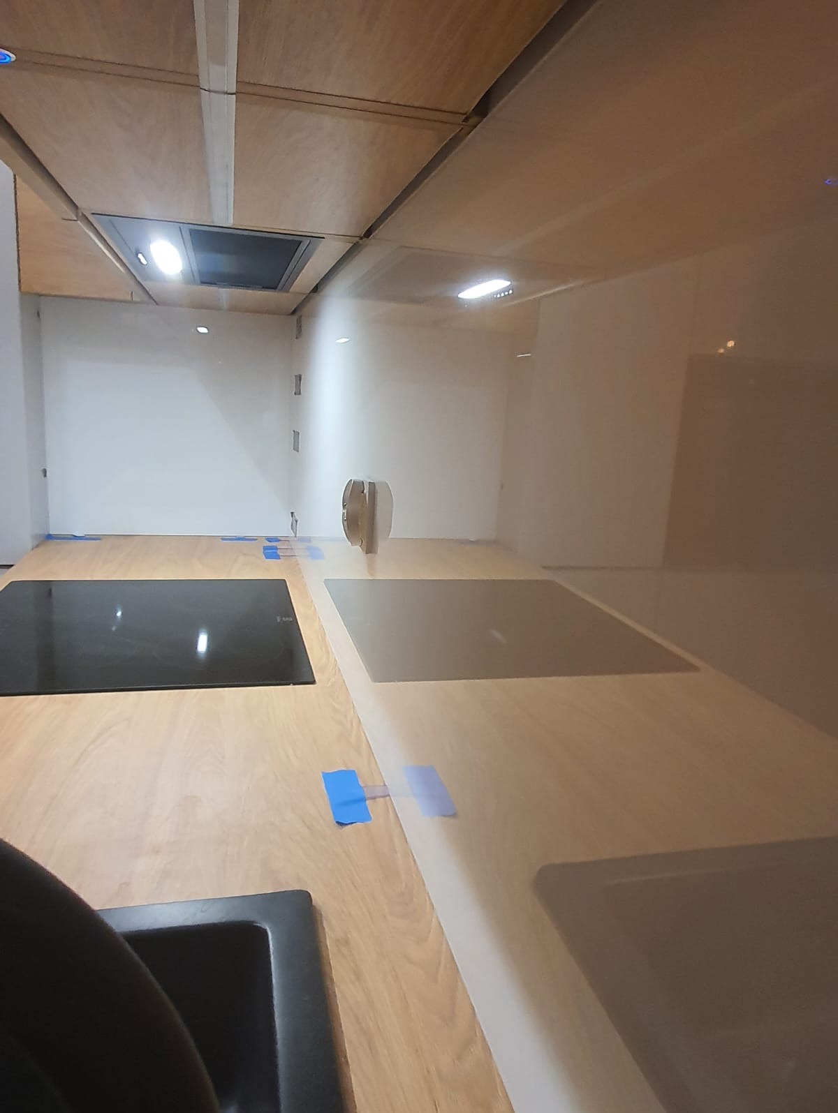 Szklana ściana nad blatem kuchennym z zamontowanymi płytami indukcyjnymi, zabezpieczona niebieską taśmą malarską, widok na wnętrze kuchni z drewnianymi szafkami.