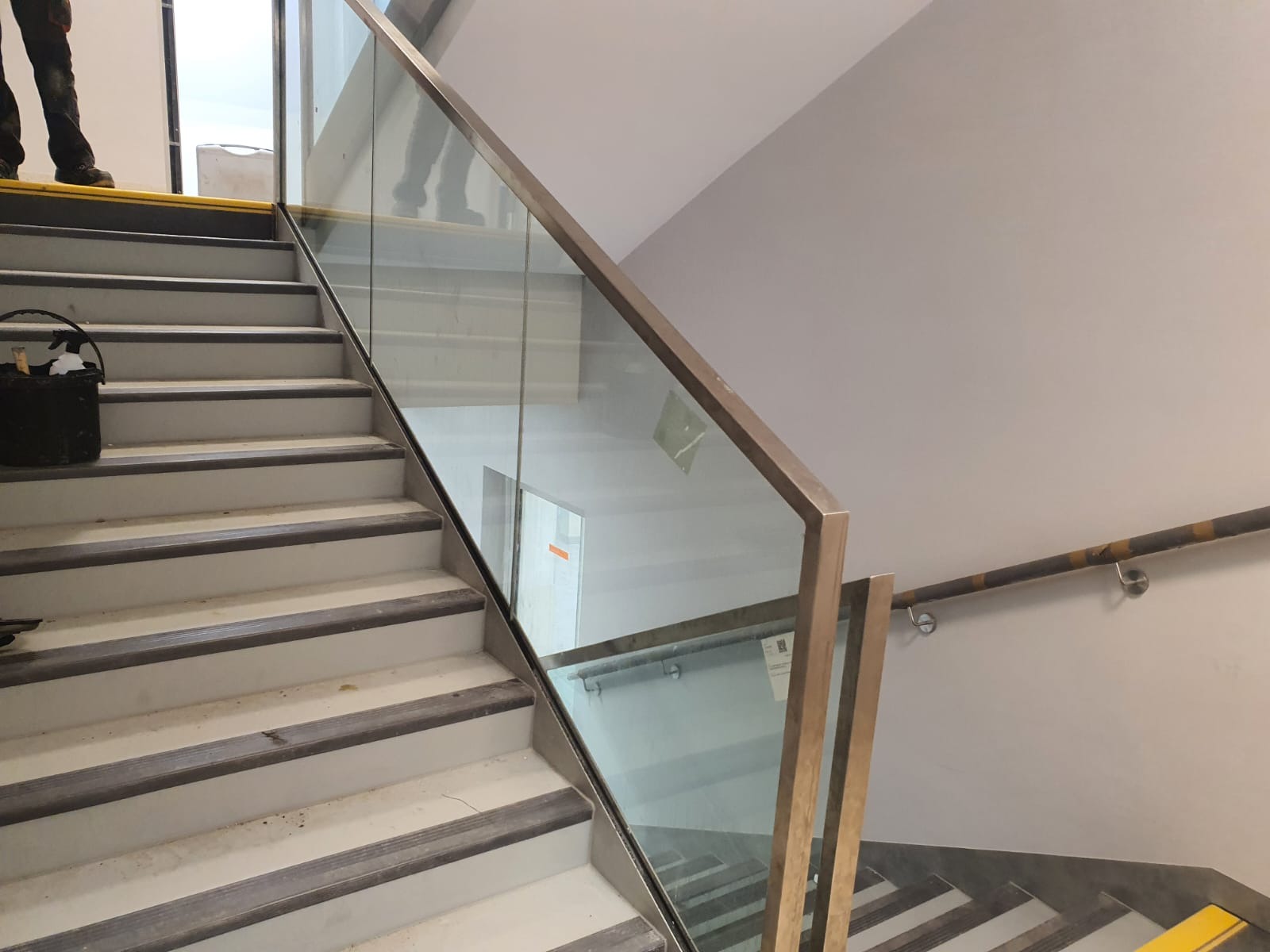 Schody z zamontowaną szklaną balustradą ze stalową ramą, widoczny kubeł z narzędziami na stopniach, częściowo widoczna osoba na schodach.
