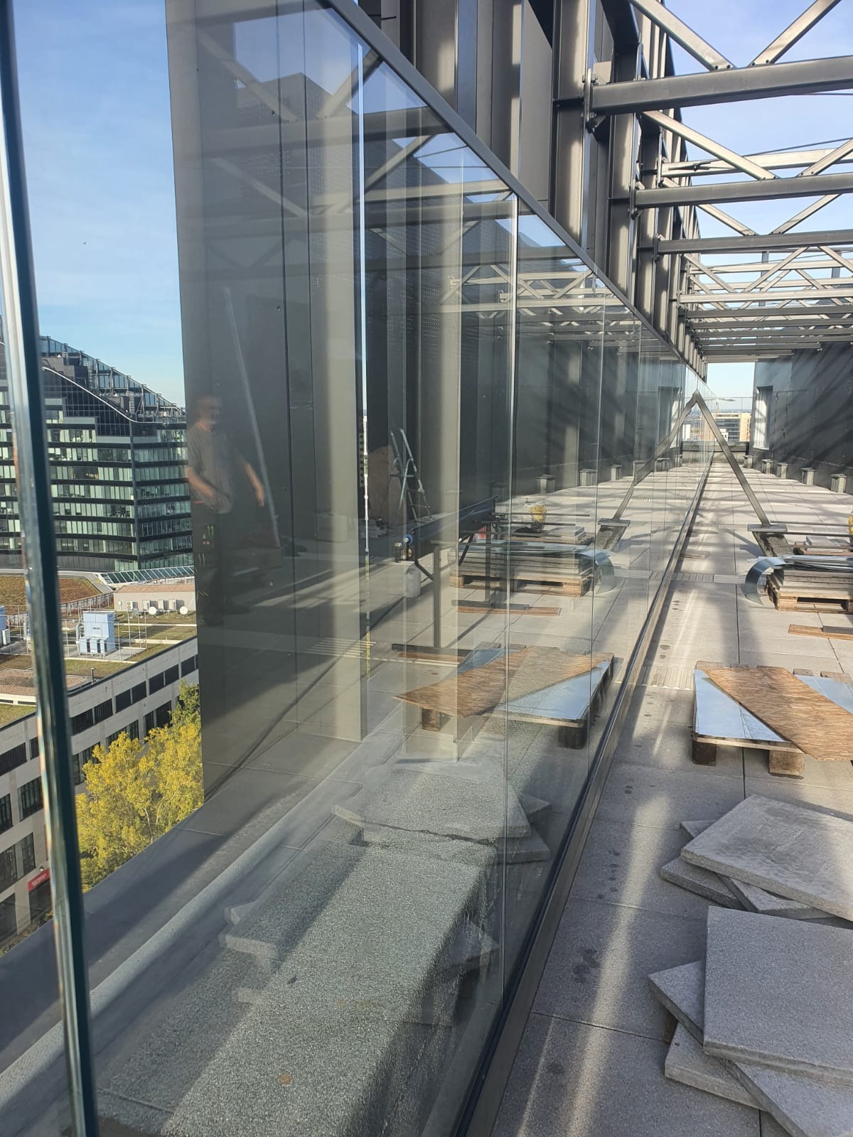 Montaż szklanej balustrady na tarasie budynku, widoczne płyty chodnikowe i narzędzia pracy, w tle zabudowa miejska.