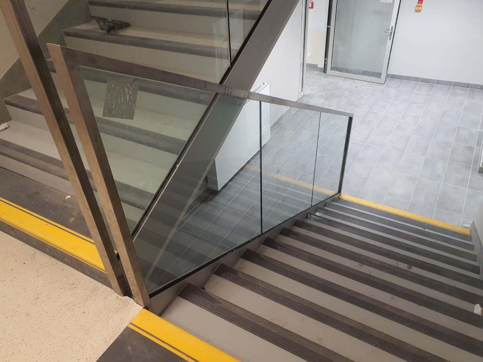 Szklana balustrada schodowa z metalowymi słupkami, widok z góry na schody z antypoślizgowymi paskami, w tle jasne płytki podłogowe i drzwi.
