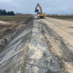 Żółto-biała koparka Liebherr 906 na placu budowy, wyrównująca teren pod drogę, widoczny świeżo ułożony nasyp z ziemi i kamienia, w tle drzewa i pochmurne niebo.