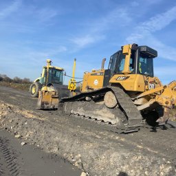 Diggertruck - Żółty spychacz gąsienicowy CAT oraz żółta ładowarka kołowa AMMANN na placu budowy, widoczne świeże ślady gąsienic i opon na ubitej ziemi, błękitne niebo w tle.
