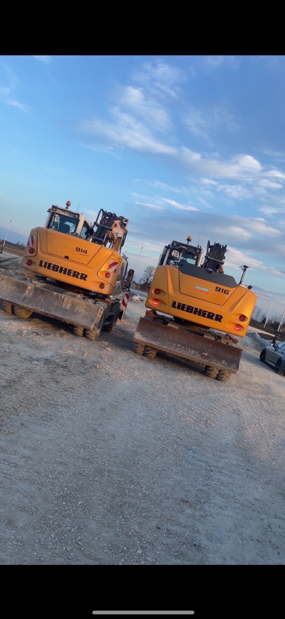 Dwa żółte, kołowe koparki Liebherr 914 i 916, ustawione tyłem do kamery na żwirowym placu budowy, widoczne elementy konstrukcyjne w tle i błękitne niebo z chmurami.