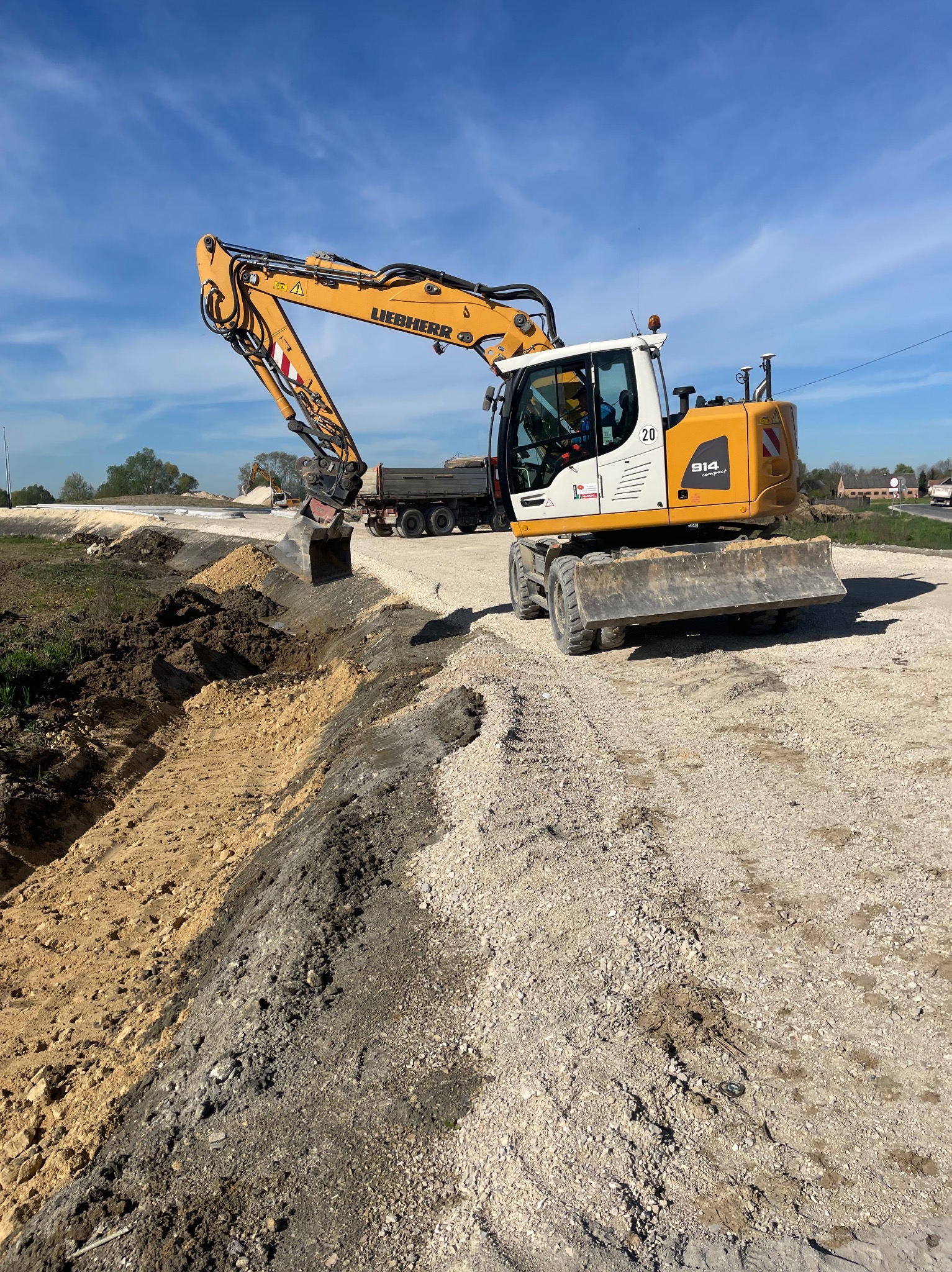 Żółta koparka kołowa Liebherr 914 Compact podczas prac ziemnych, z wykopanym rowem i ciężarówką w tle na placu budowy w słoneczny dzień.