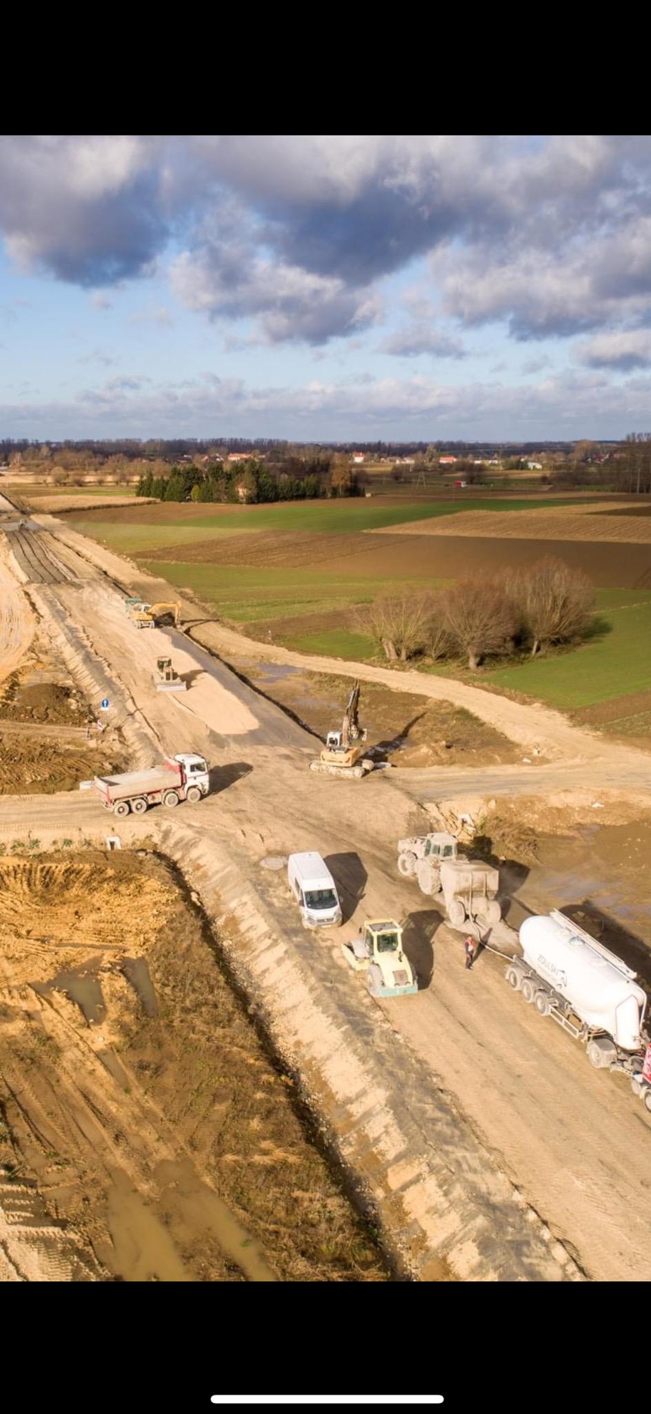Szeroki kadr z lotu ptaka ukazuje plac budowy drogi z pracującymi maszynami, w tym walcem, koparką i ciężarówką, na tle krajobrazu pól i drzew.