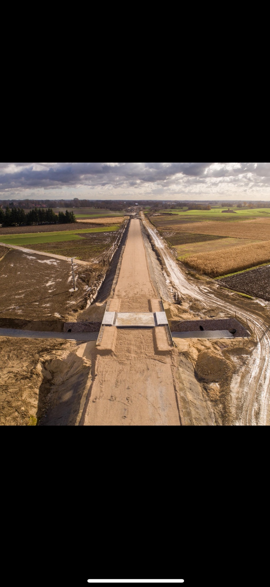 Widok z lotu ptaka na nowo budowaną drogę gruntową przez pola uprawne, z widocznym przepustem pod drogą oraz przygotowanymi rowami odwadniającymi po bokach, w pochmurny dzień.