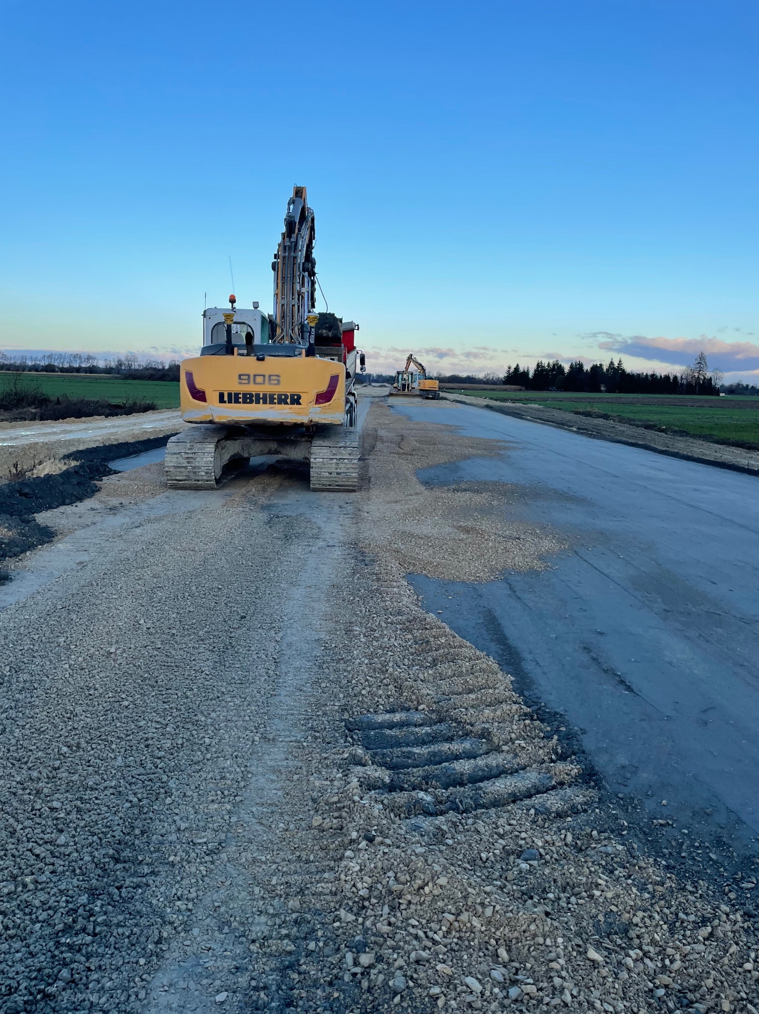 Żółta koparka Liebherr 906 na nowo budowanej drodze, z widocznymi śladami gąsienic na żwirowym podłożu i innym sprzętem budowlanym w tle, pod błękitnym niebem o zmierzchu.