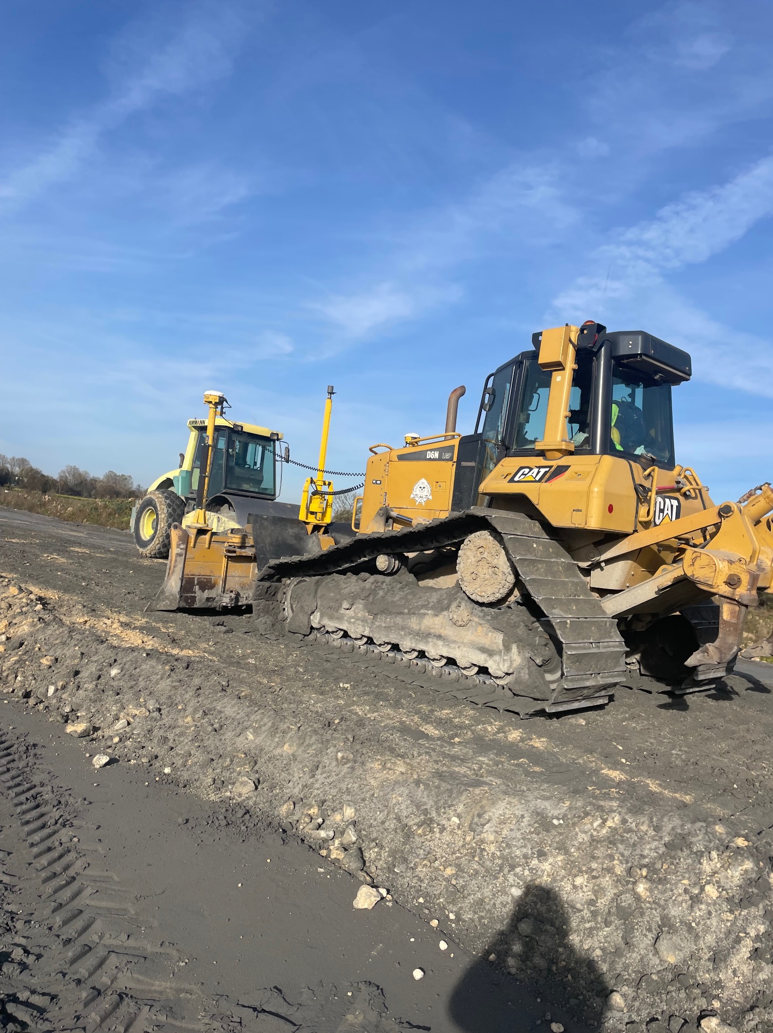 Żółty spychacz gąsienicowy CAT oraz żółta ładowarka kołowa AMMANN na placu budowy, widoczne świeże ślady gąsienic i opon na ubitej ziemi, błękitne niebo w tle.