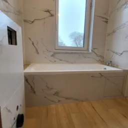 Wykończona łazienka z wanną obudowaną płytkami imitującymi marmur, drewnopodobna podłoga, okno z widokiem na drzewa.