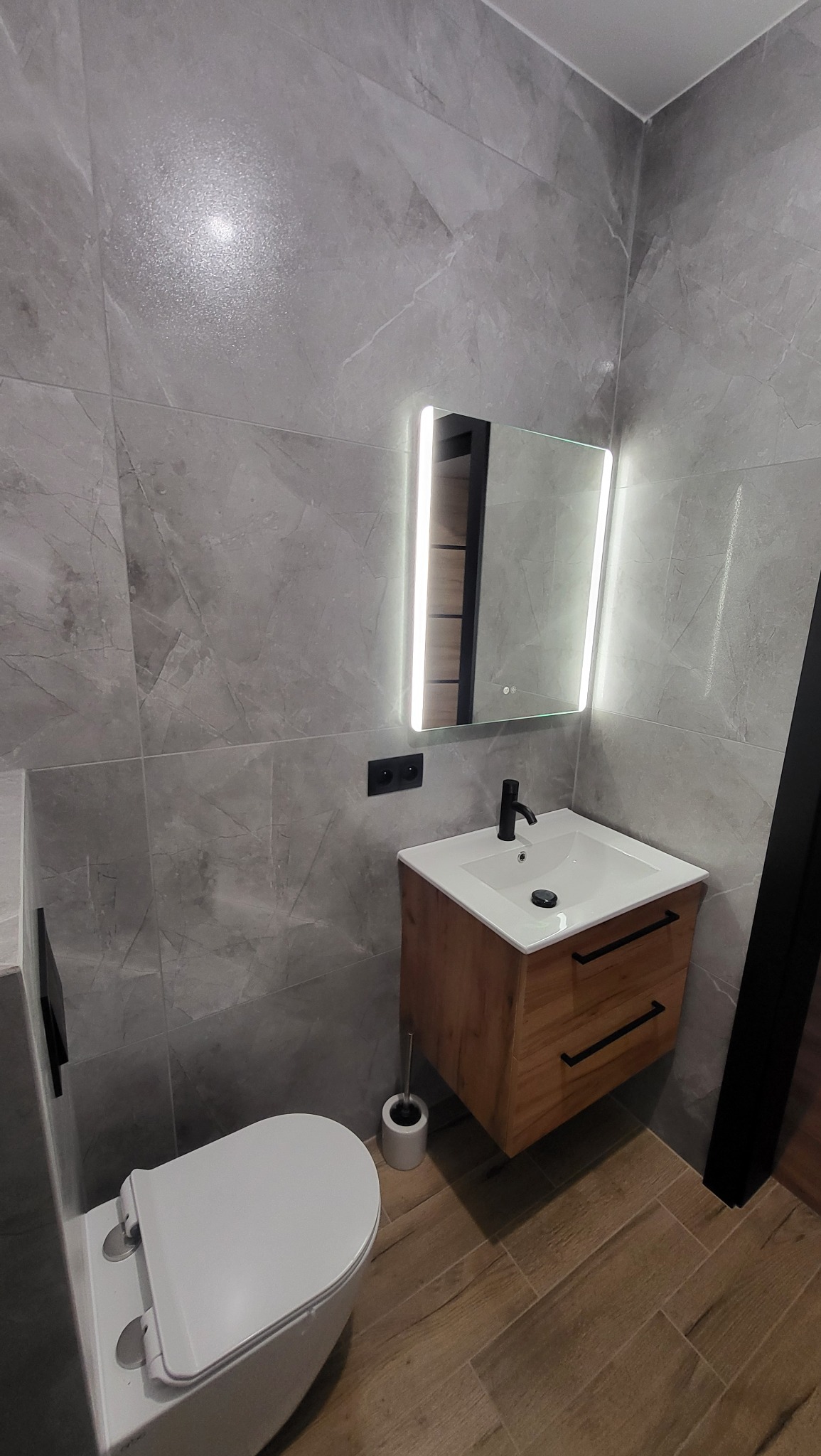 Nowoczesna łazienka z szarymi płytkami imitującymi marmur, podświetlanym lustrem nad drewnianą szafką z umywalką, białą toaletą i drewnopodobną podłogą.