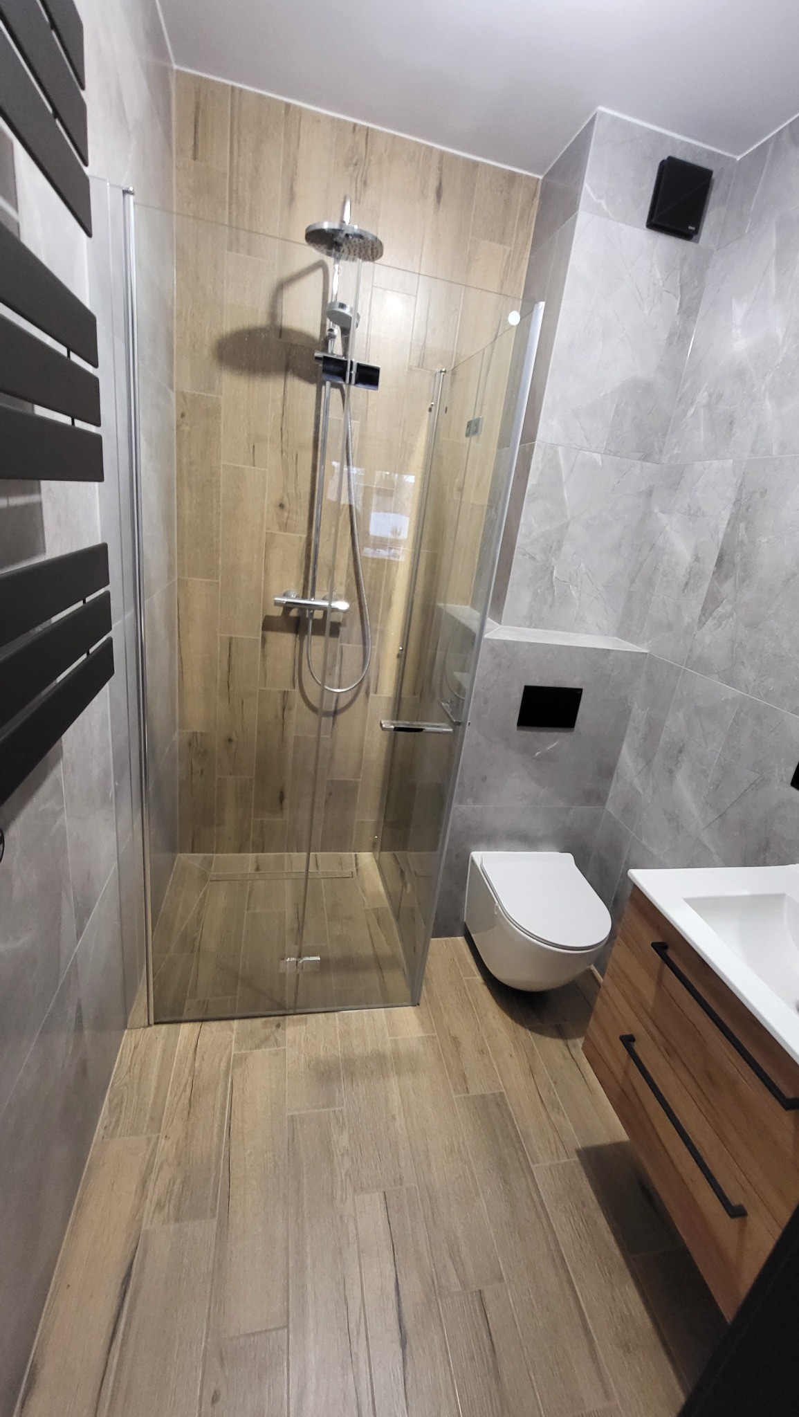 Nowoczesna łazienka z kabiną prysznicową typu walk-in, wykończona płytkami imitującymi drewno i szary marmur, podwieszana toaleta z czarnym przyciskiem spłukiwania i szafka pod umywalkę...