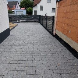 Piotr Salachna - Ułożona szara kostka brukowa na podjeździe, z widocznym ogrodzeniem panelowym i fragmentem budynku w trakcie budowy w tle.